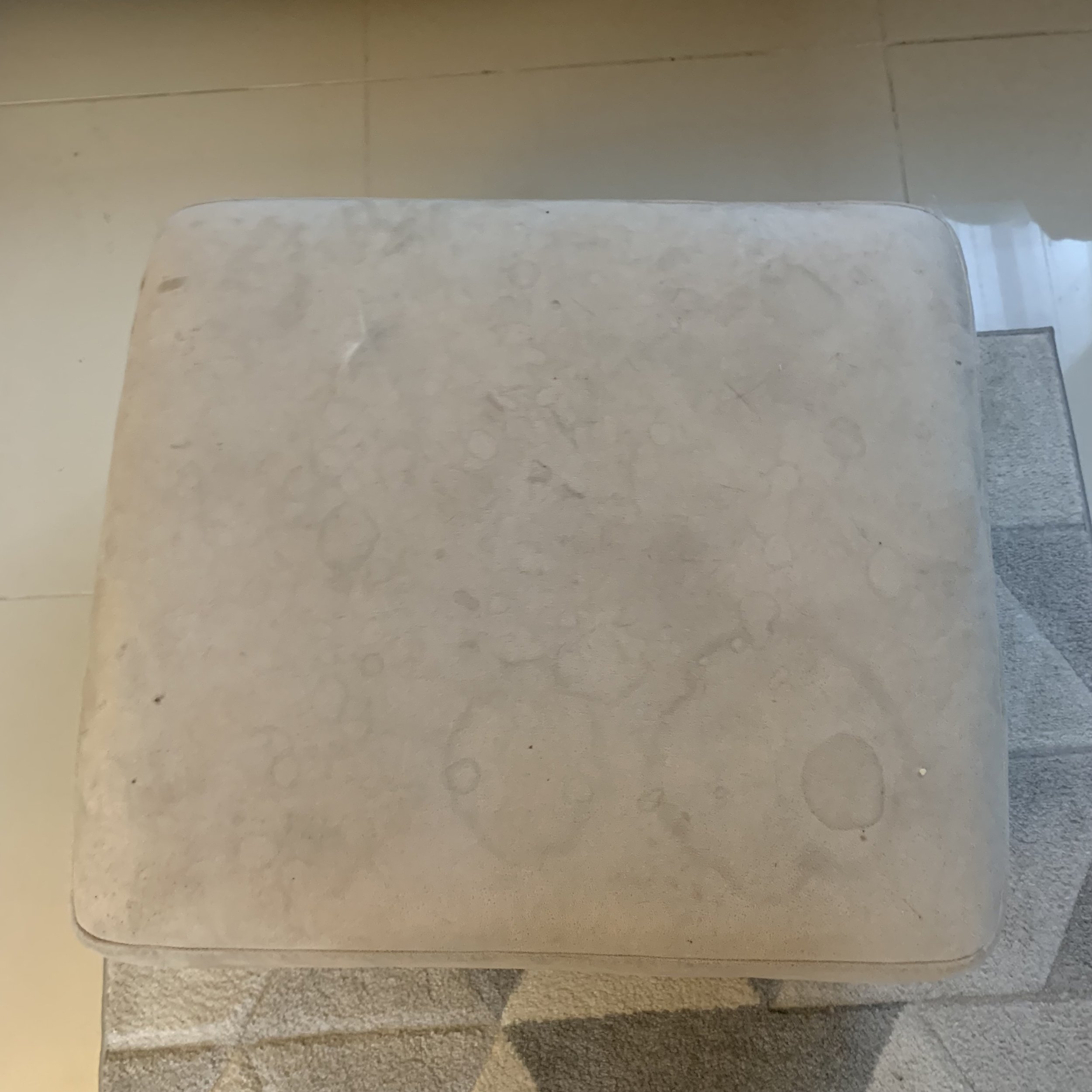 A beige marble top table with a smooth surface and rounded edges, placed on a tiled floor with a textured rug partially visible underneath.