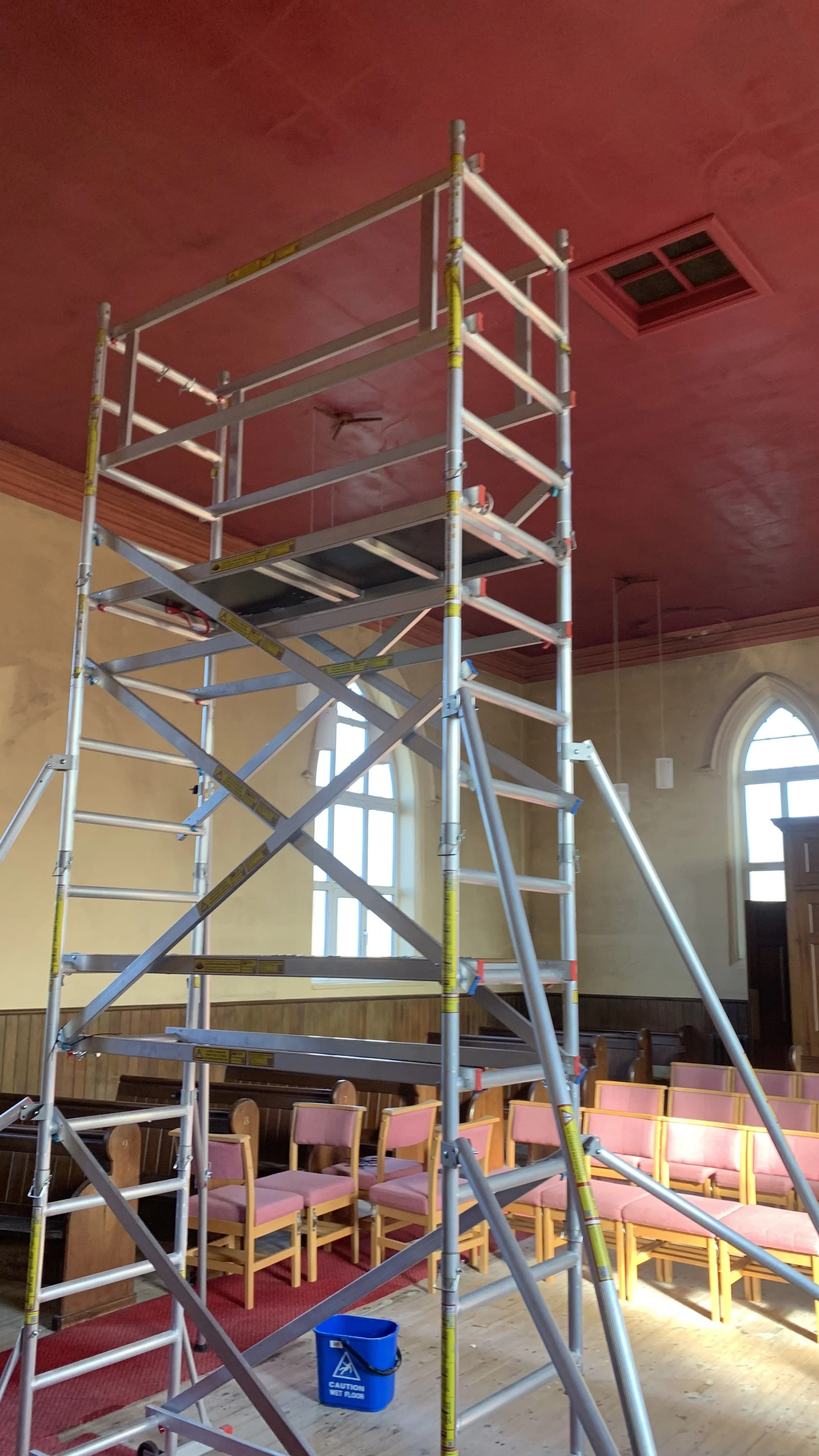 A tall aluminum scaffolding structure inside a room with pink ceiling and wooden chairs along the walls.