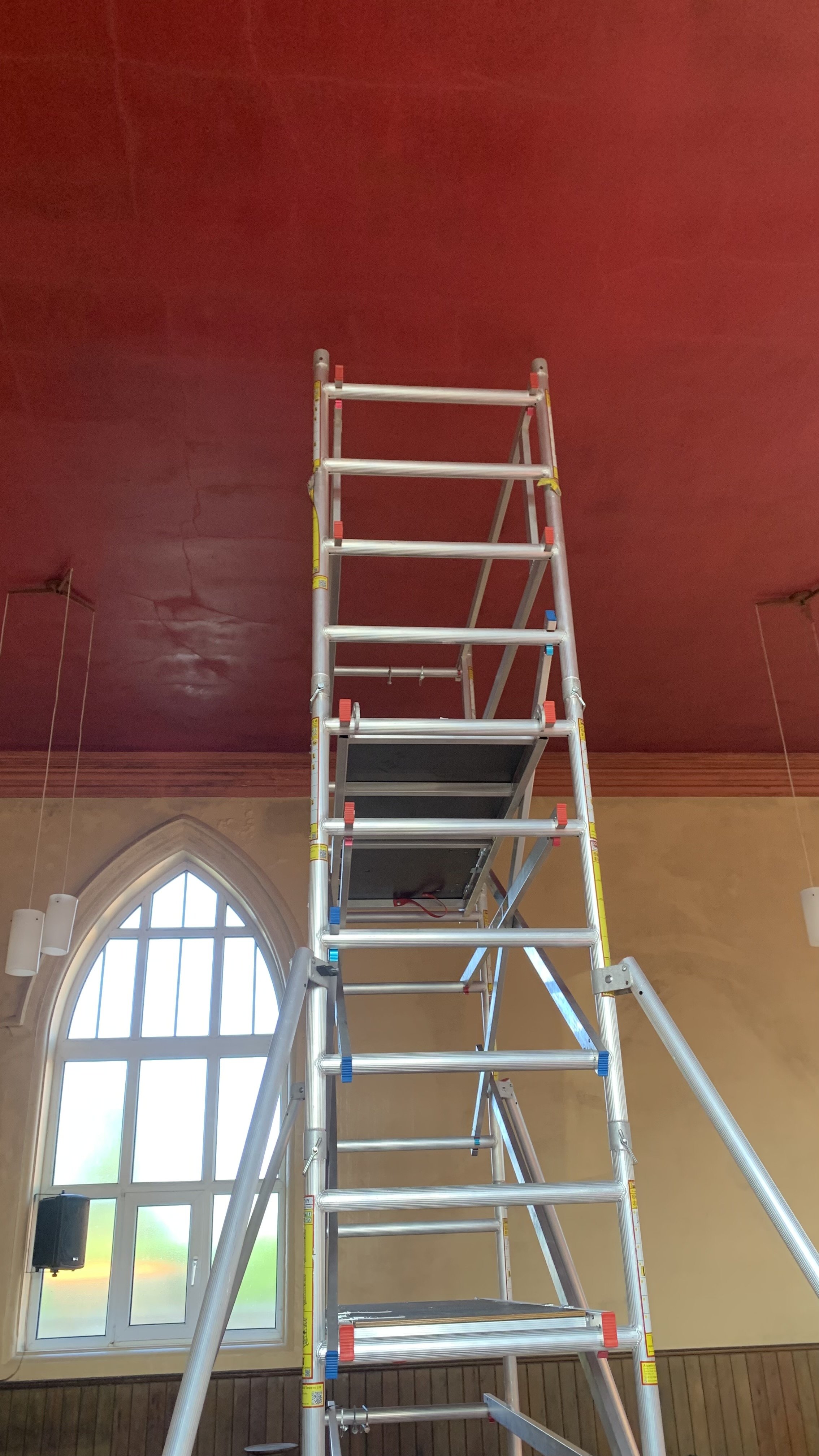 A tall aluminum ladder extending towards a red ceiling in a room with yellow walls and a large arched window.