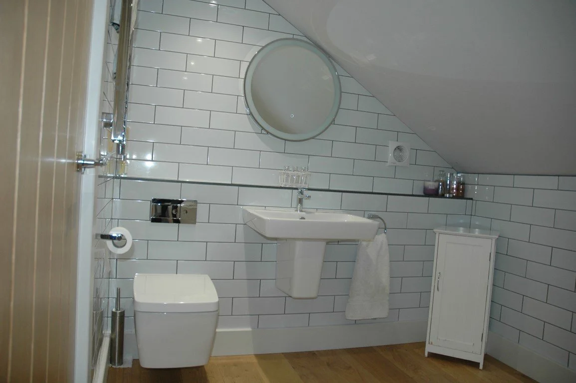 Bathroom with white tile walls, a wall-mounted sink with a towel hanging underneath, a modern toilet, and a small white cabinet. There is a mirror above the sink and small decorative items on a shelf.