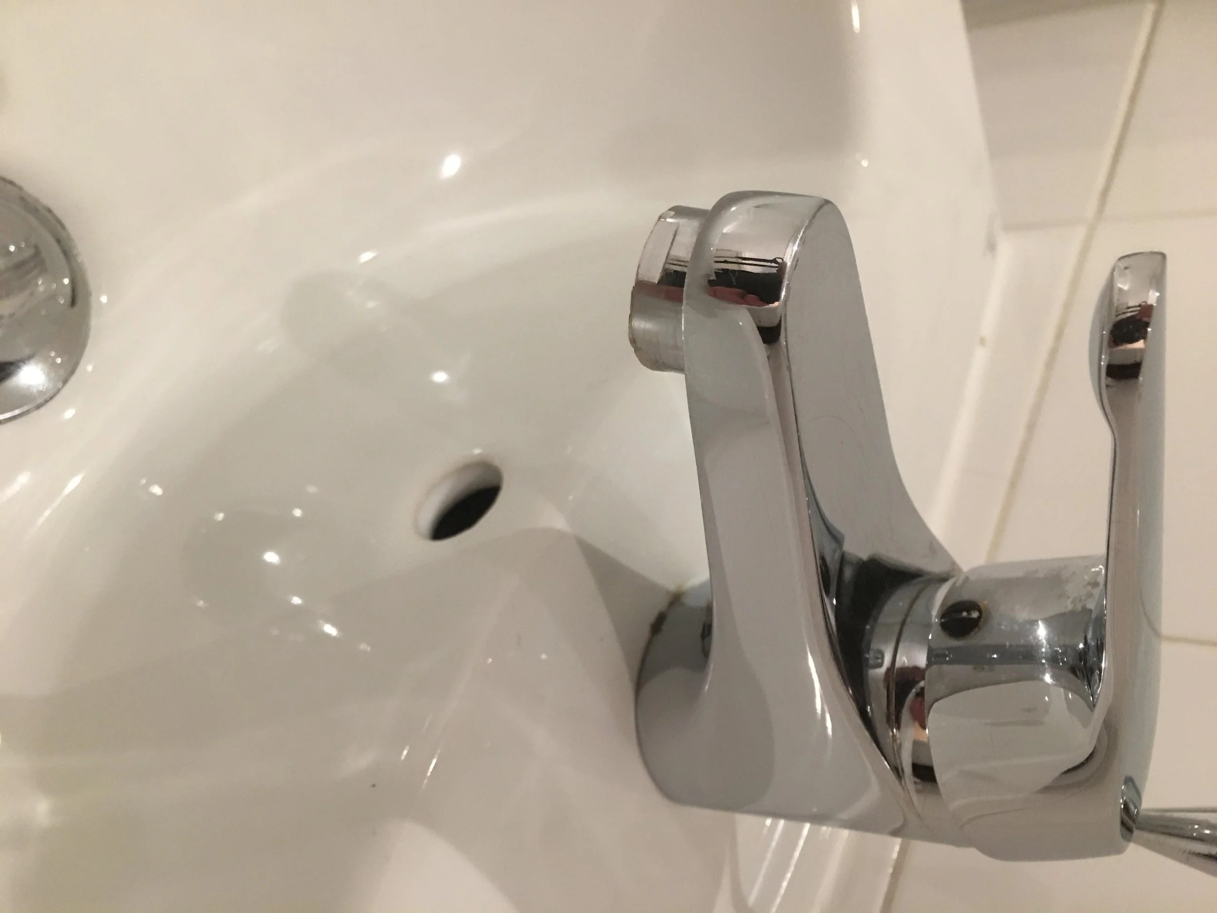 Close-up of a modern chrome bathroom faucet over a white sink with a drain and tiled wall in the background.