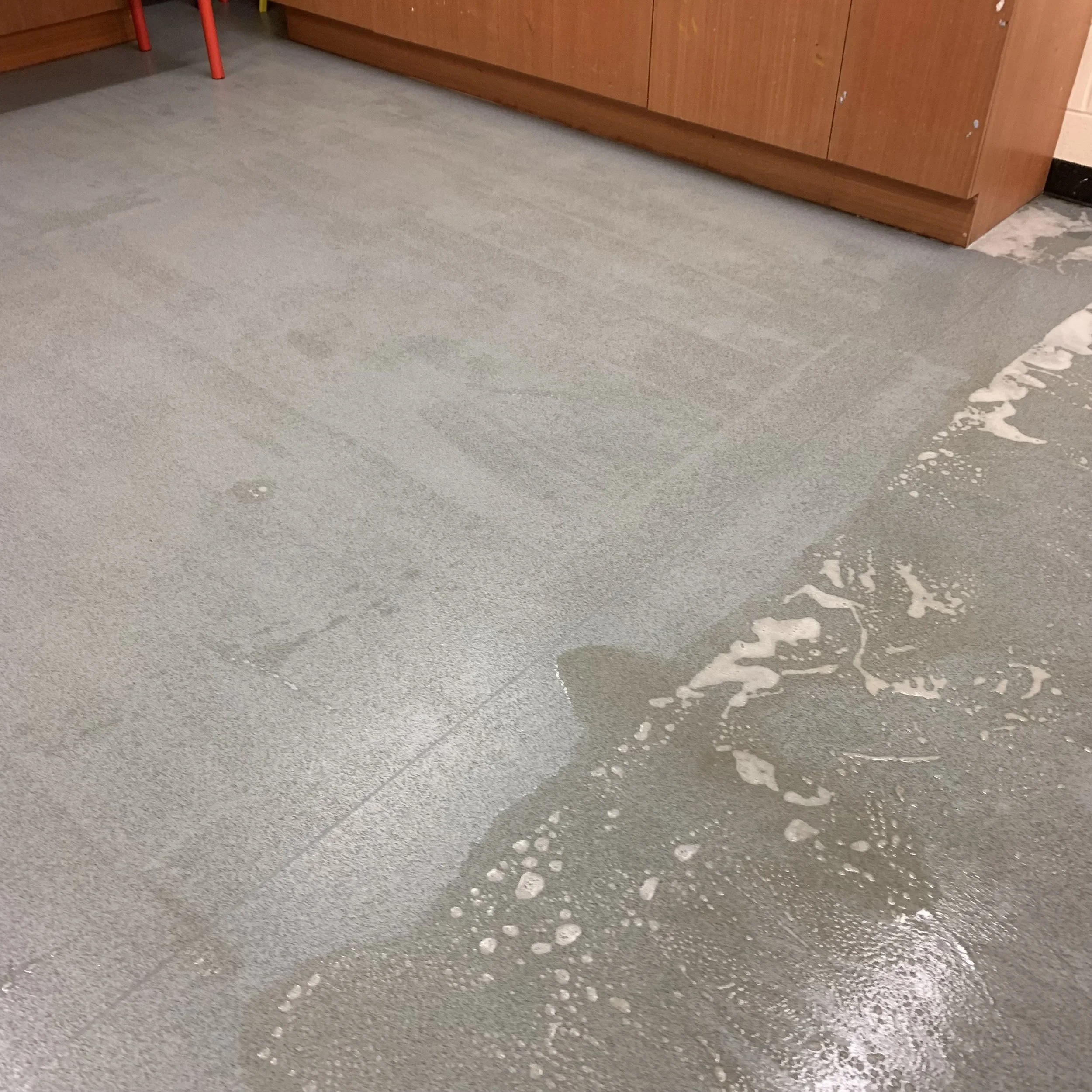 A wet floor with soapy water and foam on a gray tiled surface near a wooden cabinet.