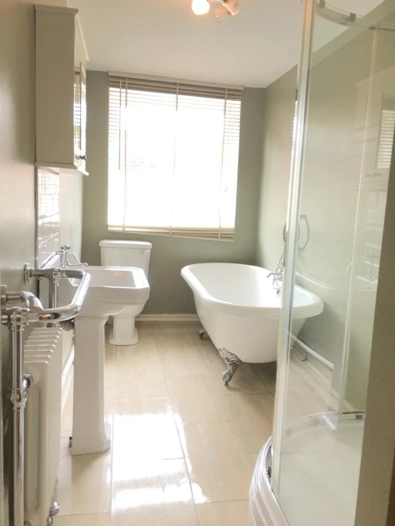 A bathroom with a window, a clawfoot bathtub, a toilet, a pedestal sink, and a curved standing shower, with beige tiled floor and green walls.