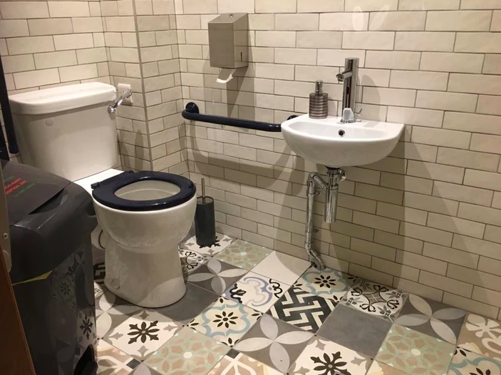 A small accessible bathroom with a toilet, small wall-mounted sink, and patterned tile floor. A black grab bar is attached to the wall near the toilet.