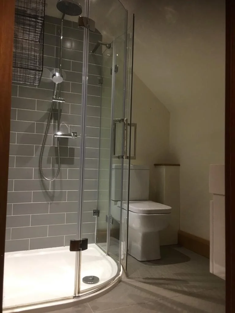 A small bathroom with a corner shower with glass doors, gray subway tile walls, a rain shower head, and a handheld shower head. Next to the shower is a toilet with a lid, and a wall-mounted cabinet is visible to the right.