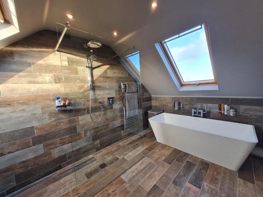 Modern bathroom with a walk-in shower with glass partition, rainfall showerhead, and tiled walls, along with a white freestanding bathtub below a skylight window, featuring wooden and earthy-toned tiles.