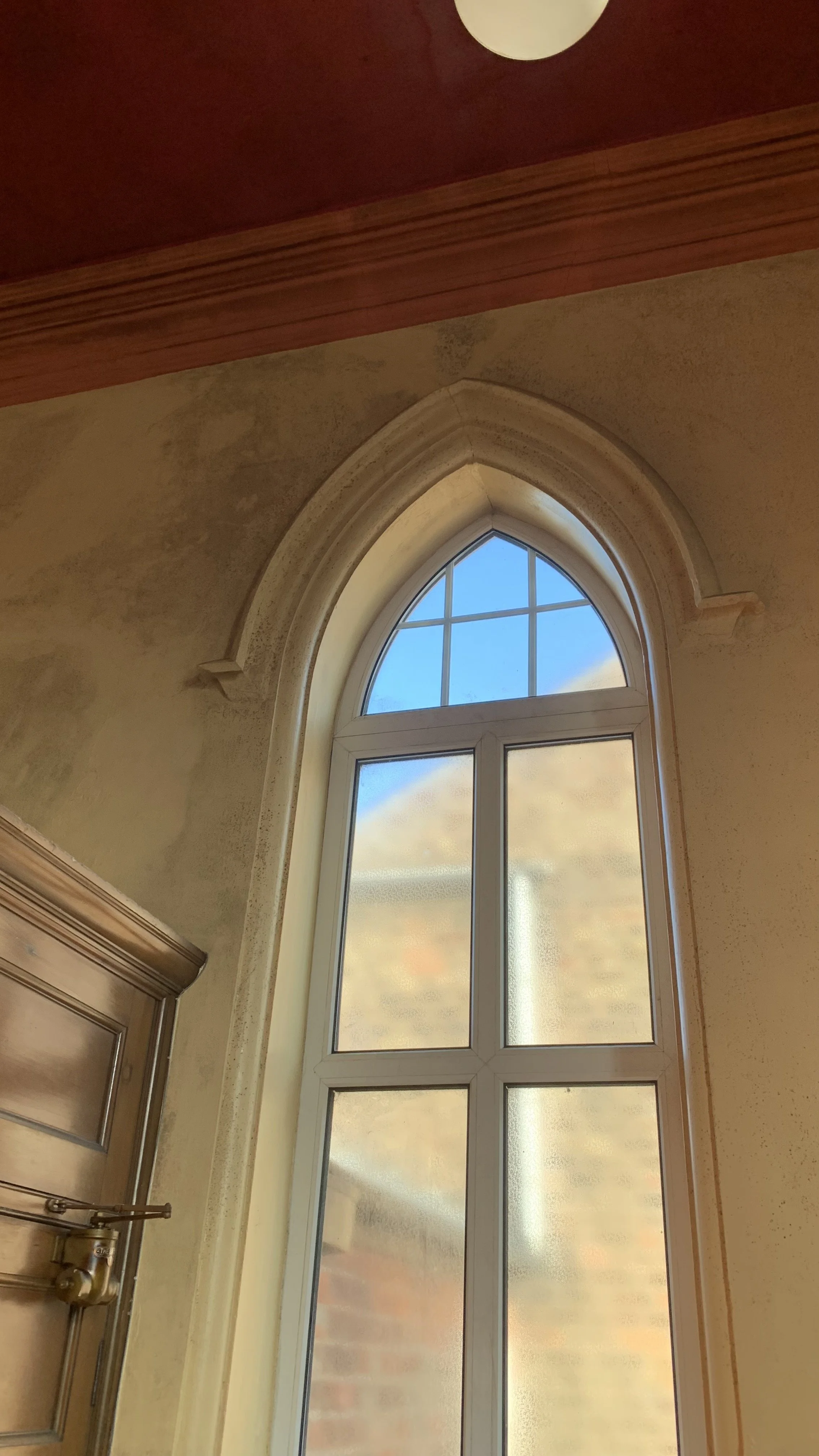 A tall arched window with multiple panes, set in a beige wall inside a building. The window shows a blue sky with some clouds outside.