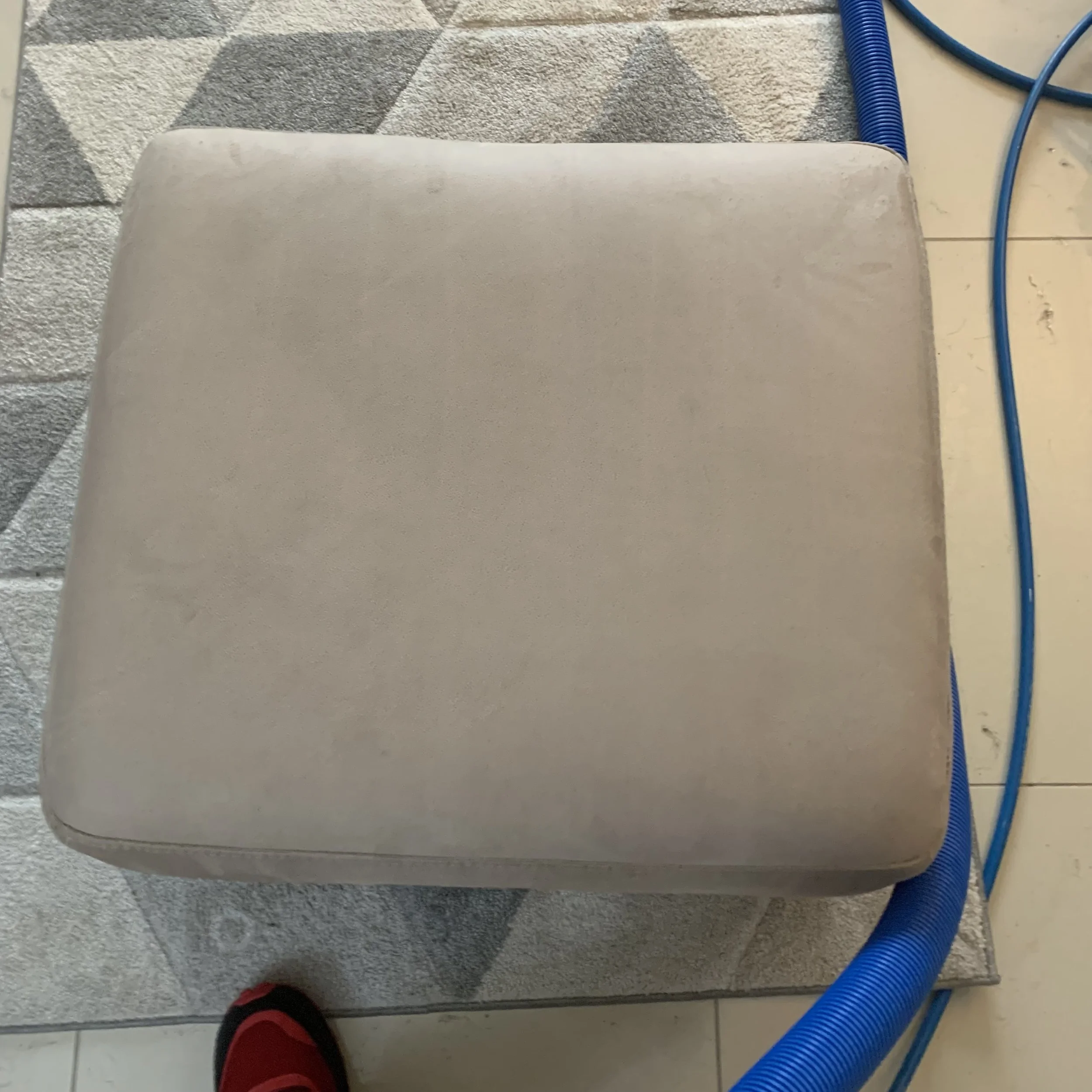 A beige cushioned stool on a geometric patterned rug, with blue hoses around it and a person's red shoe visible at the bottom.