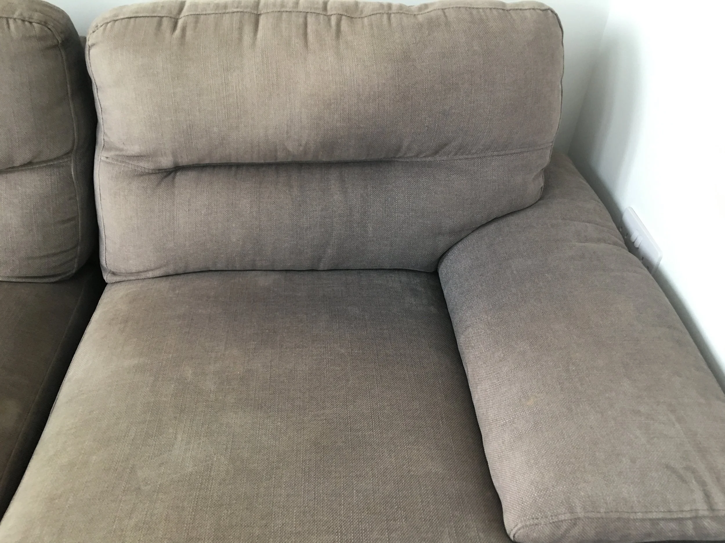 A beige fabric sectional sofa with a cushioned armrest and backrest, situated next to a wall with a visible electrical outlet.