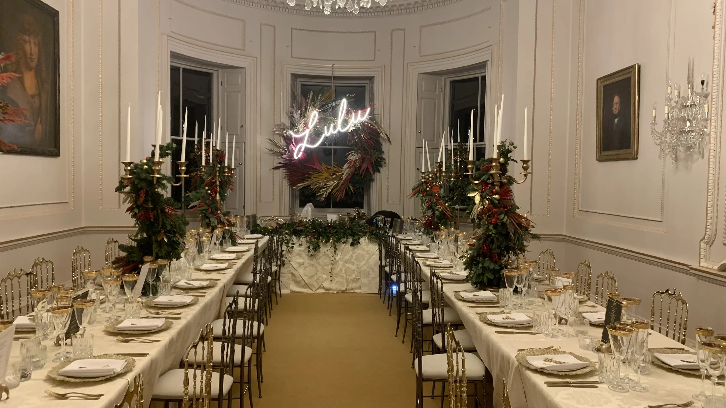 Elegant dining room decorated for Christmas with tables set with glassware, plates, and cutlery. Green garlands and red ornaments adorn gold candle holders on the tables. A floral wreath with a neon sign reading 'Love' is on the wall at the end of th