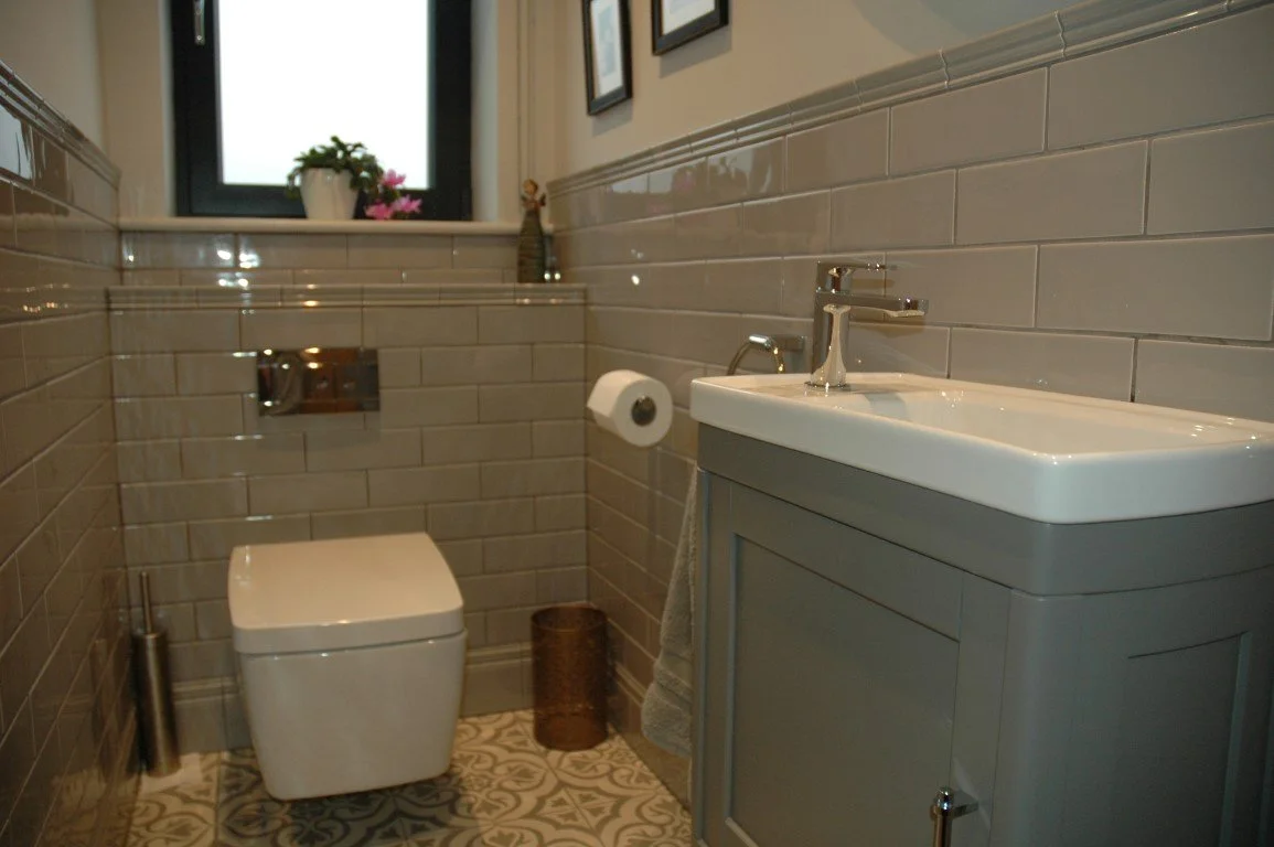 Small bathroom with a gray vanity, white sink, wall-mounted toilet, window with pink flowers, framed pictures, and beige tiled walls.
