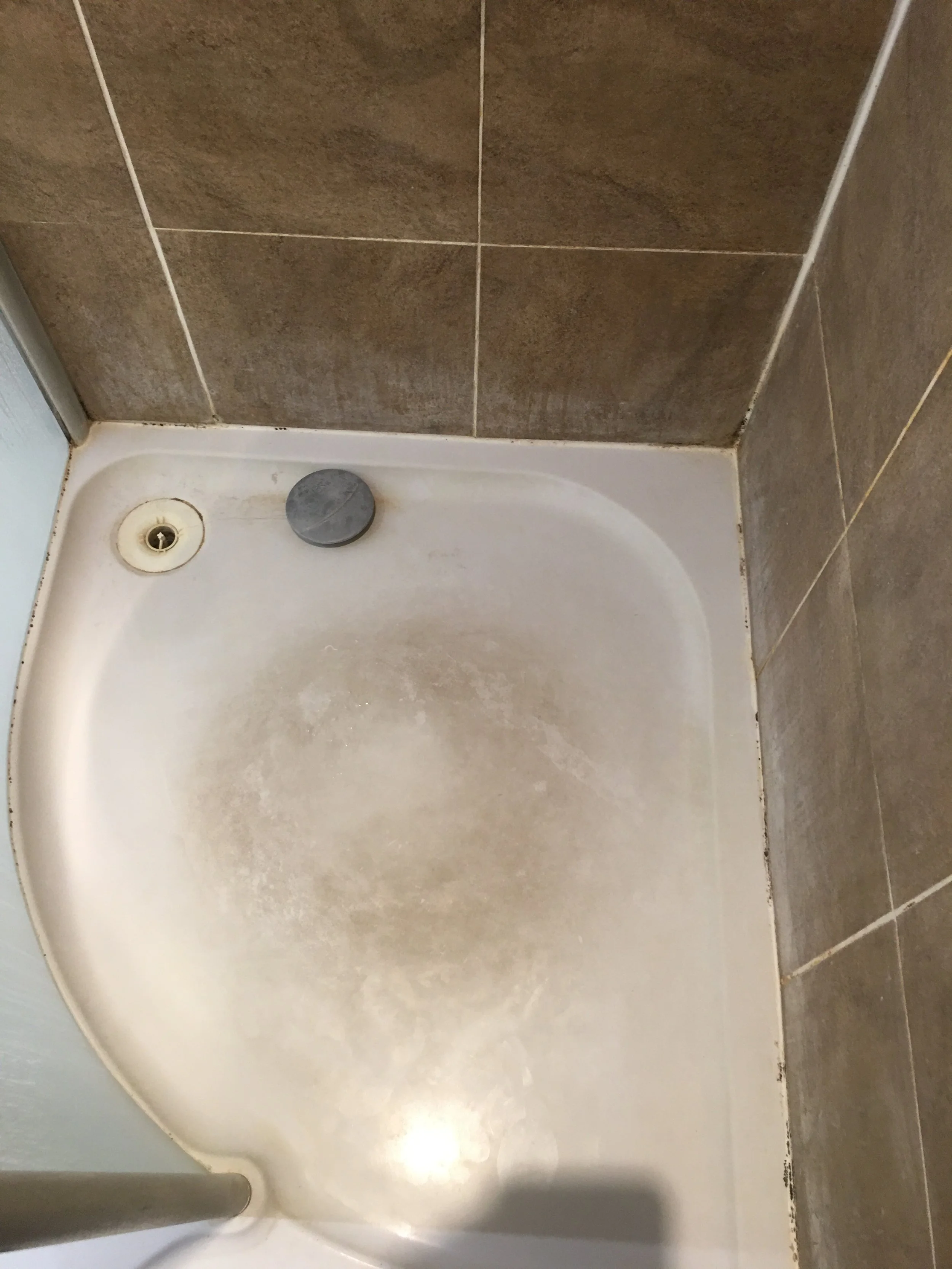 Dirty shower floor with a black drain cover and a round gray drain stopper, surrounded by brown tiled walls.