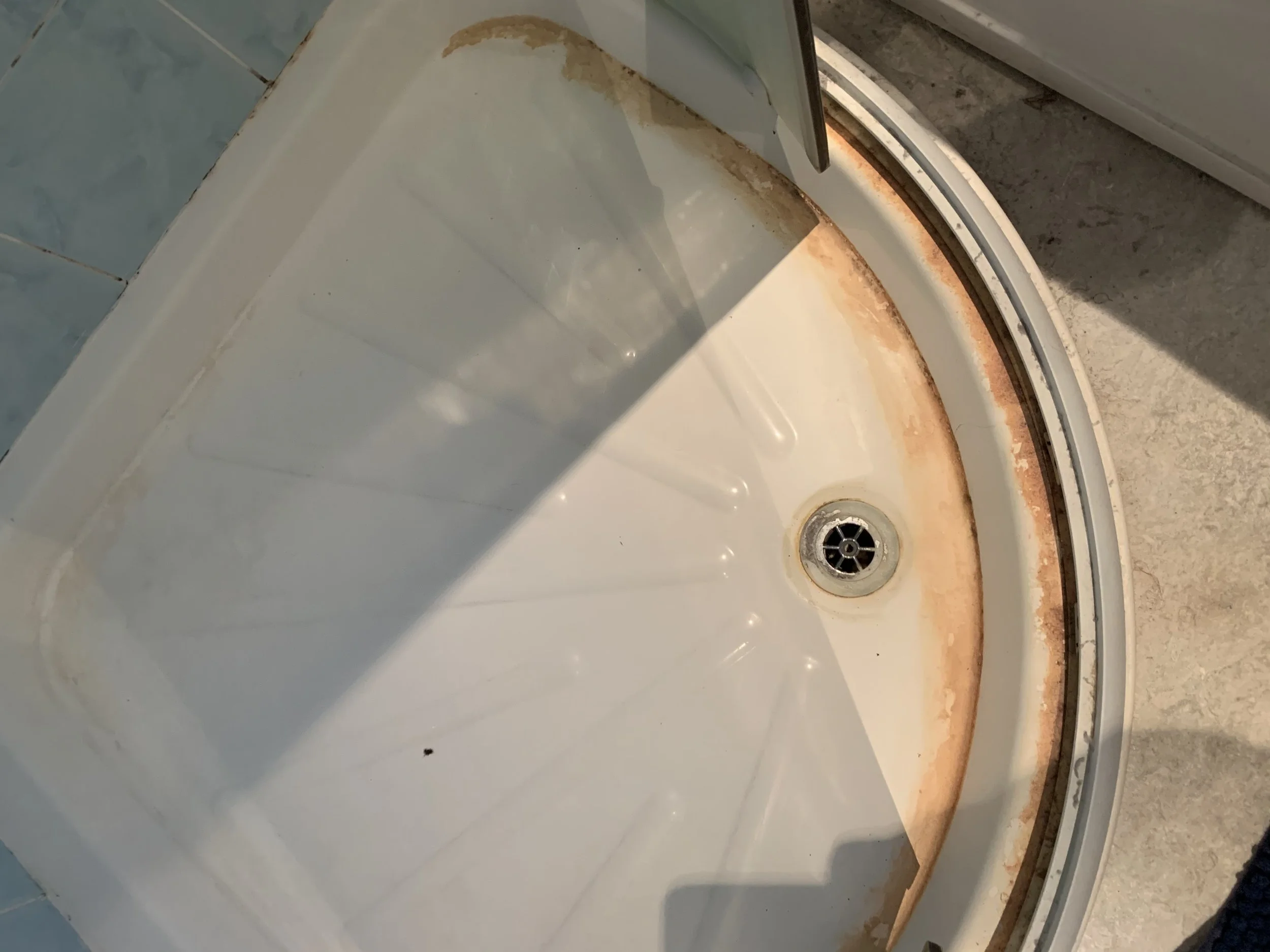 Close-up of a dirty and rusted bathtub drain with rust stains around the edge and a partially visible tiled wall.