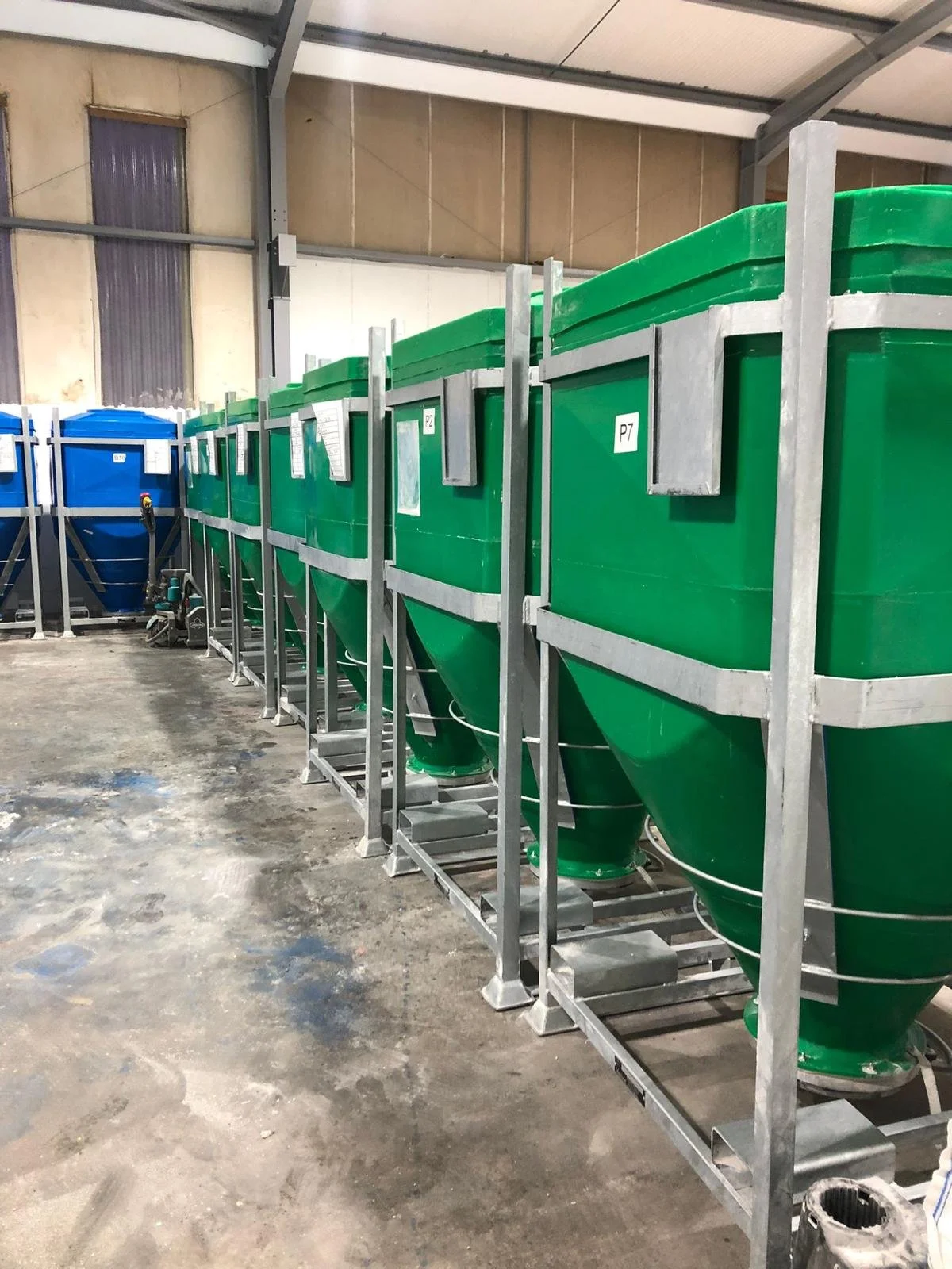 Row of large green and blue industrial hoppers or silos in a warehouse or factory setting.