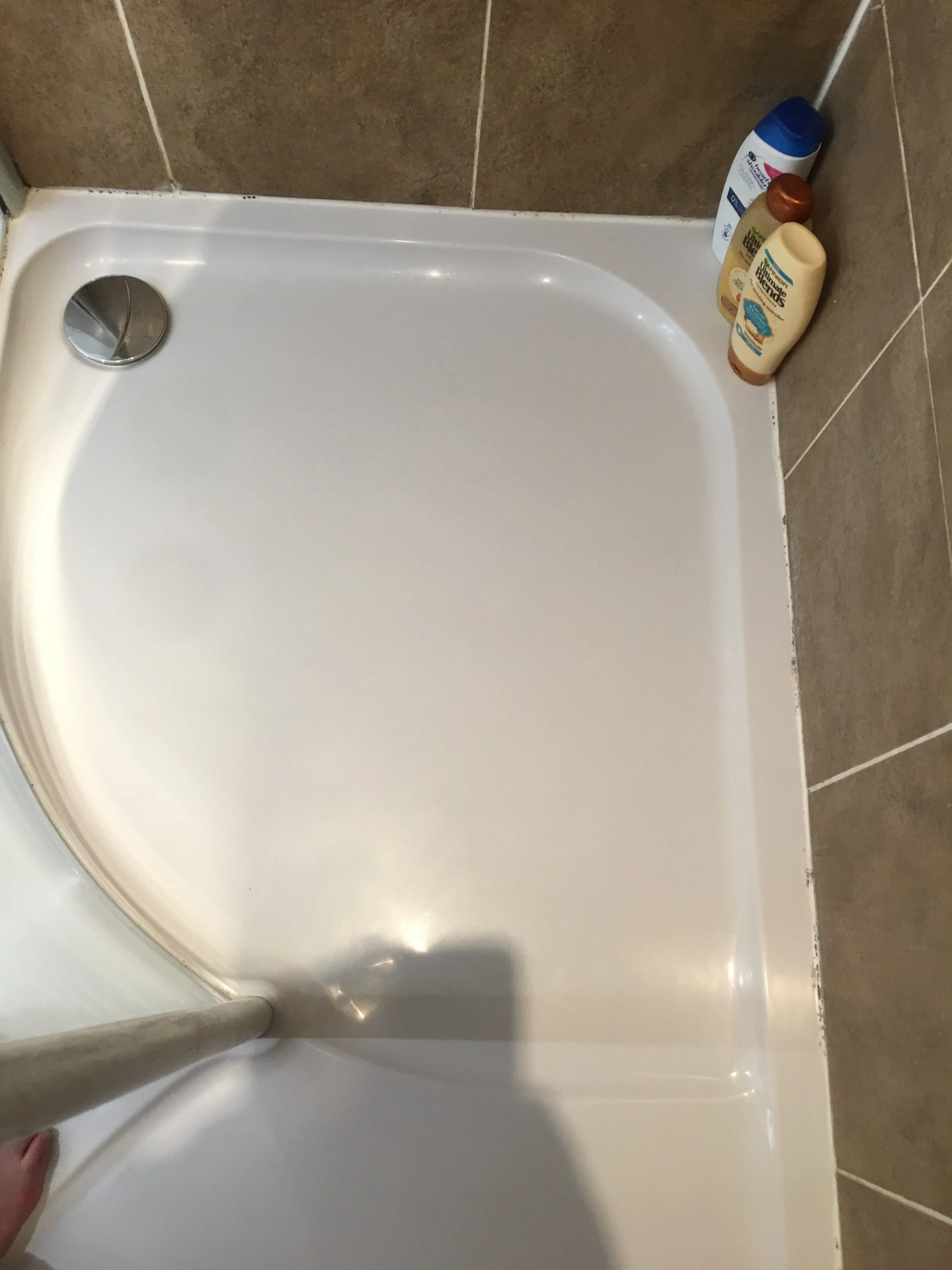 Empty white shower with a handheld shower head, brown tiled floor, and bottles of personal care products on the corner shelf.