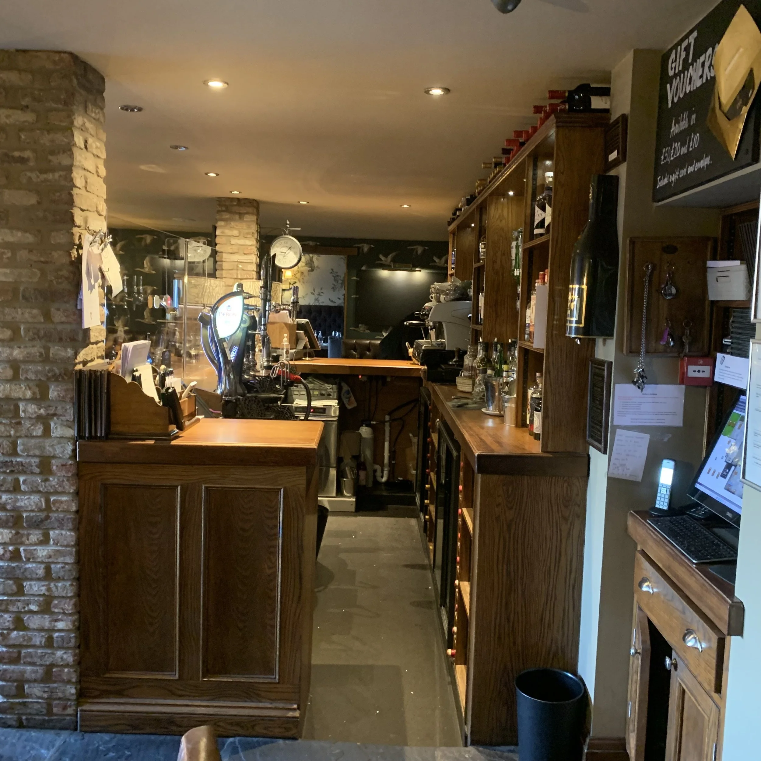 Interior of a cozy bar with wooden shelves and brick walls, featuring a counter with various drinks, coffee machines, and bar equipment, illuminated by warm small ceiling lights.