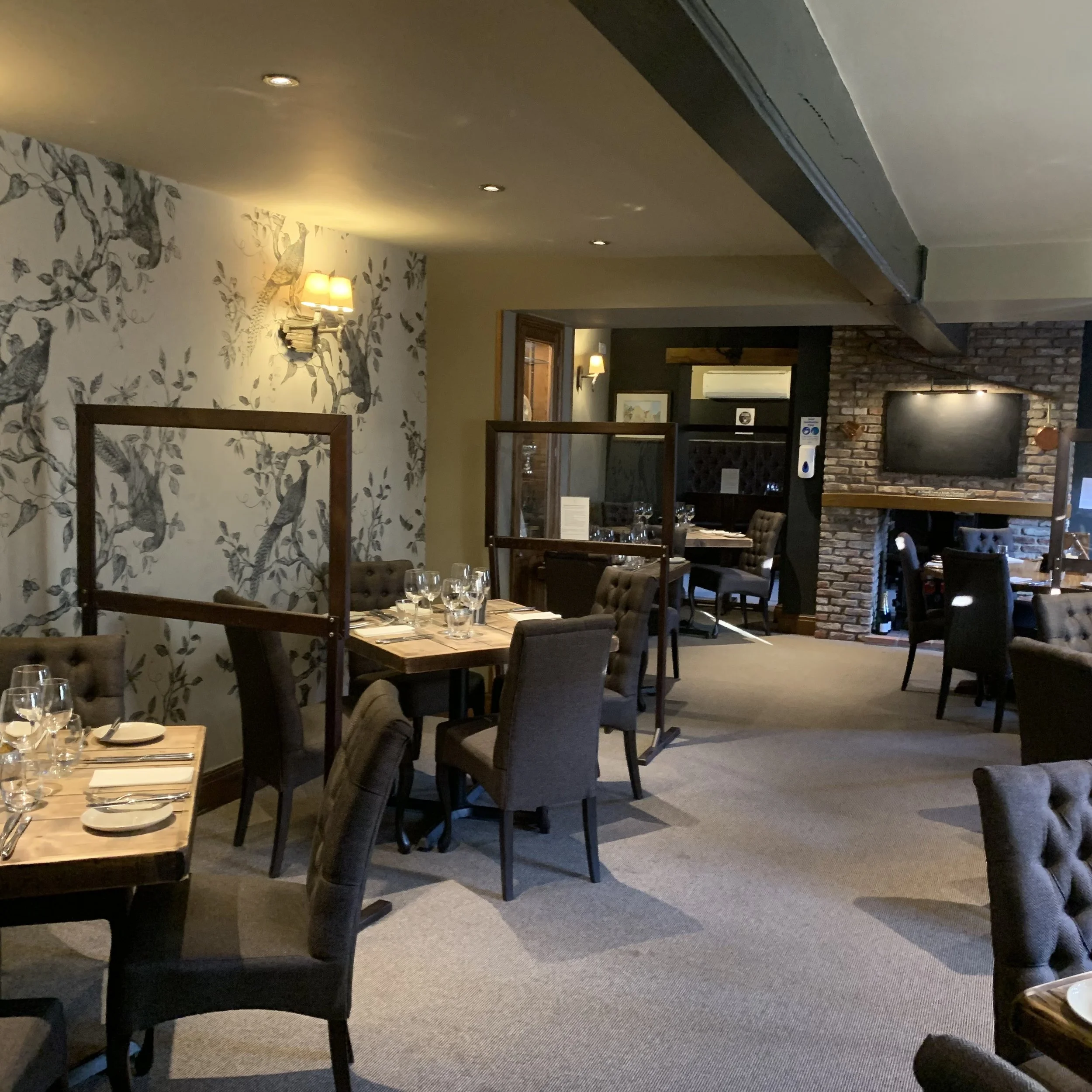 A cozy restaurant interior with wooden tables, tufted chairs, wall-mounted lamps, a brick fireplace, and decorated walls with tree and bird patterns.