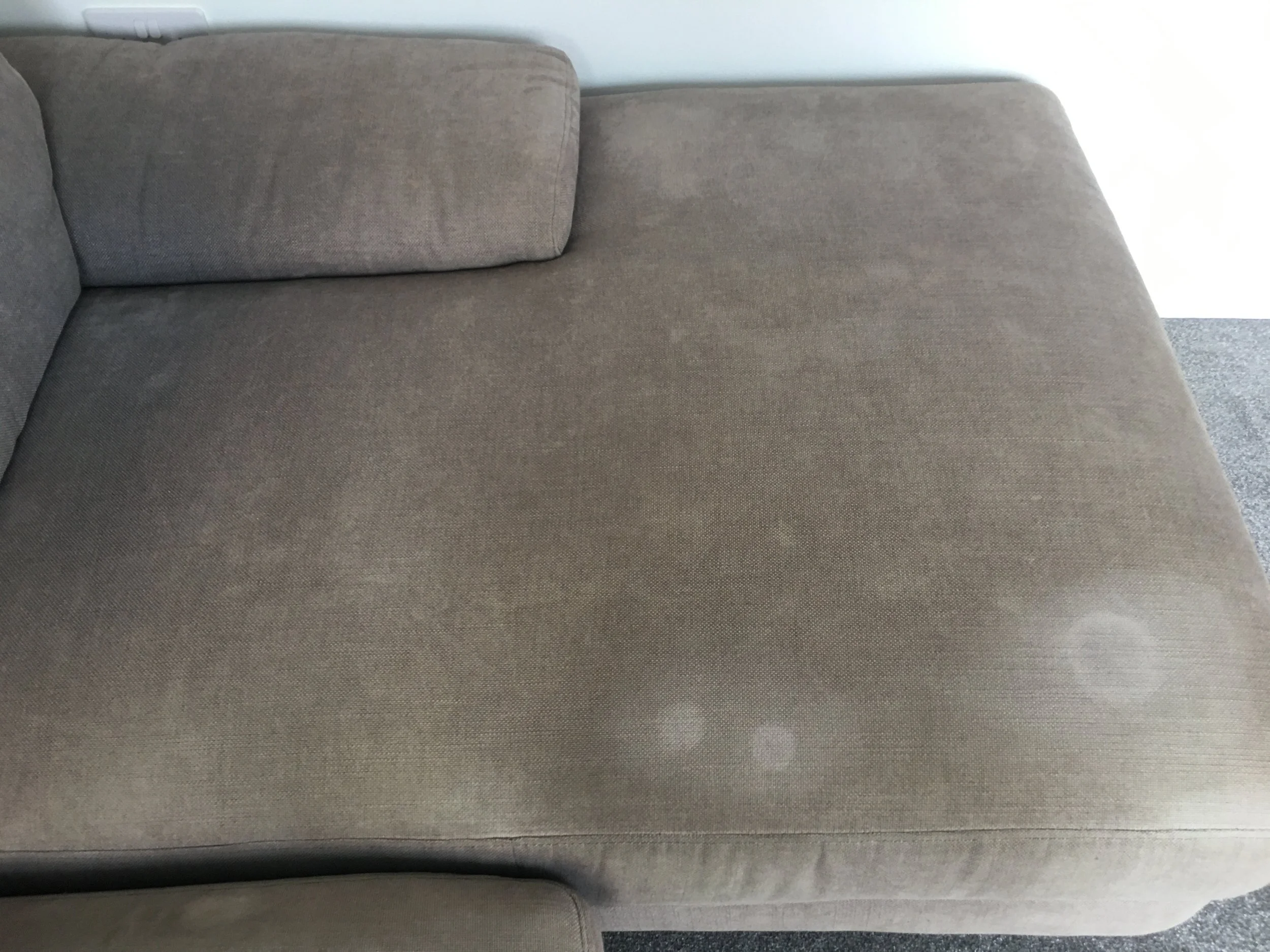 A close-up view of a beige fabric sofa with a cushion, showing some white spots and stains on the surface.