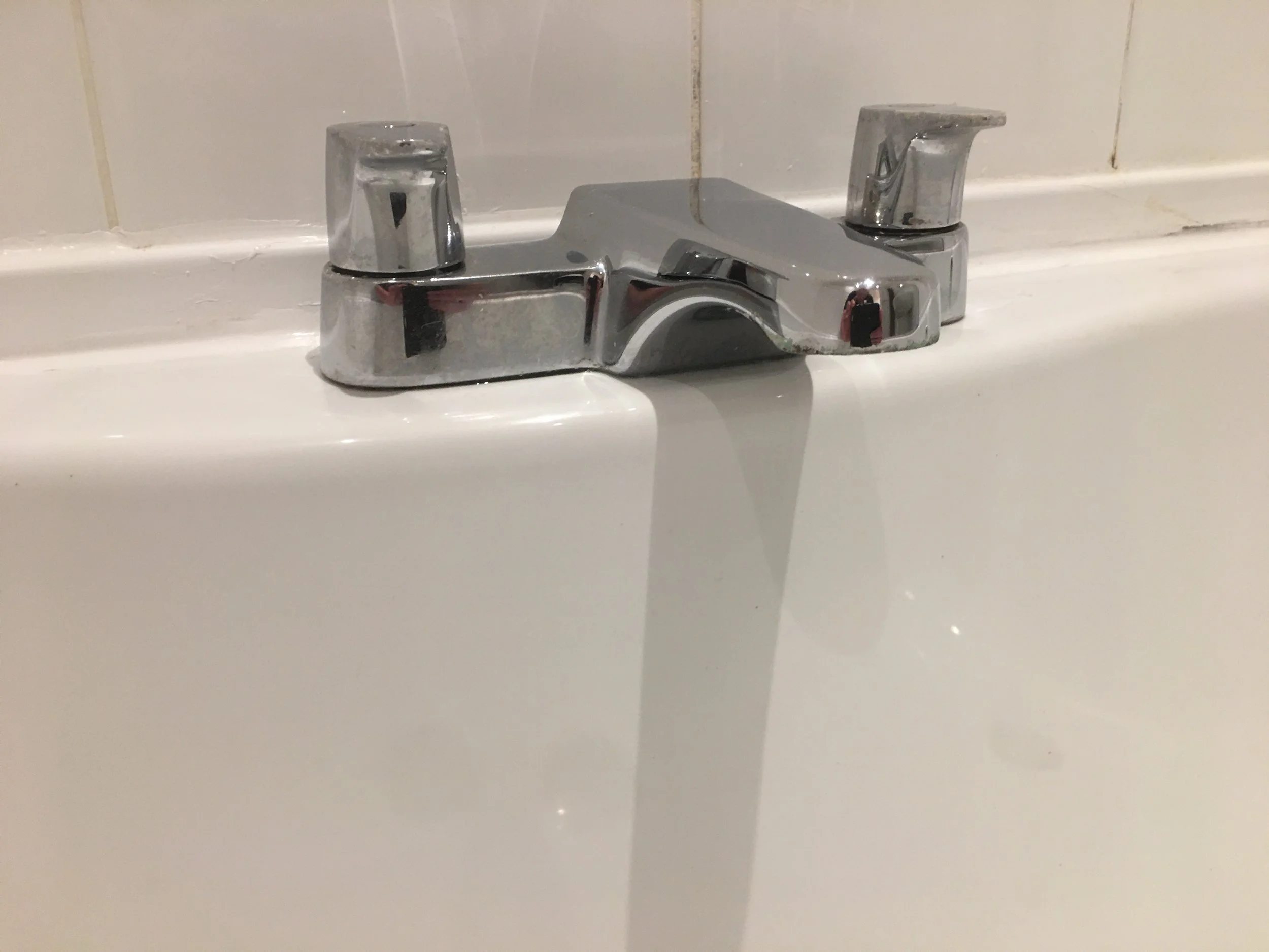 Close-up of a chrome bathroom faucet with two handles on a white sink with a tiled wall in the background.