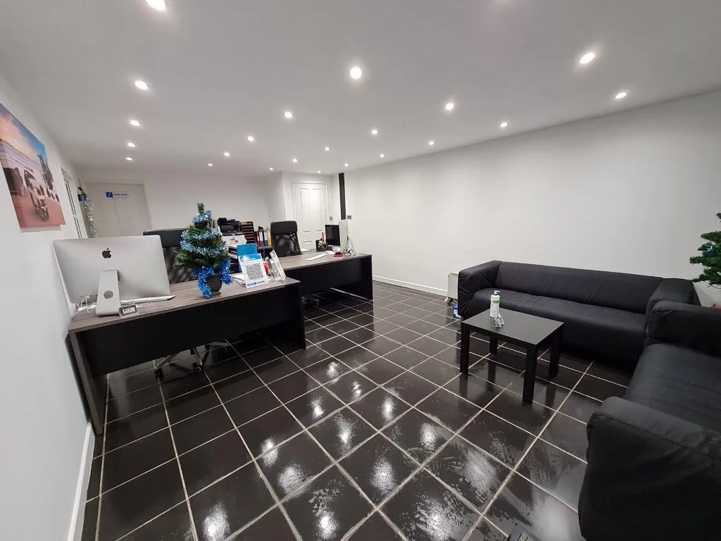 Office reception area decorated for Christmas with black leather sofas, desks with computers, a small Christmas tree, and bright ceiling lights.