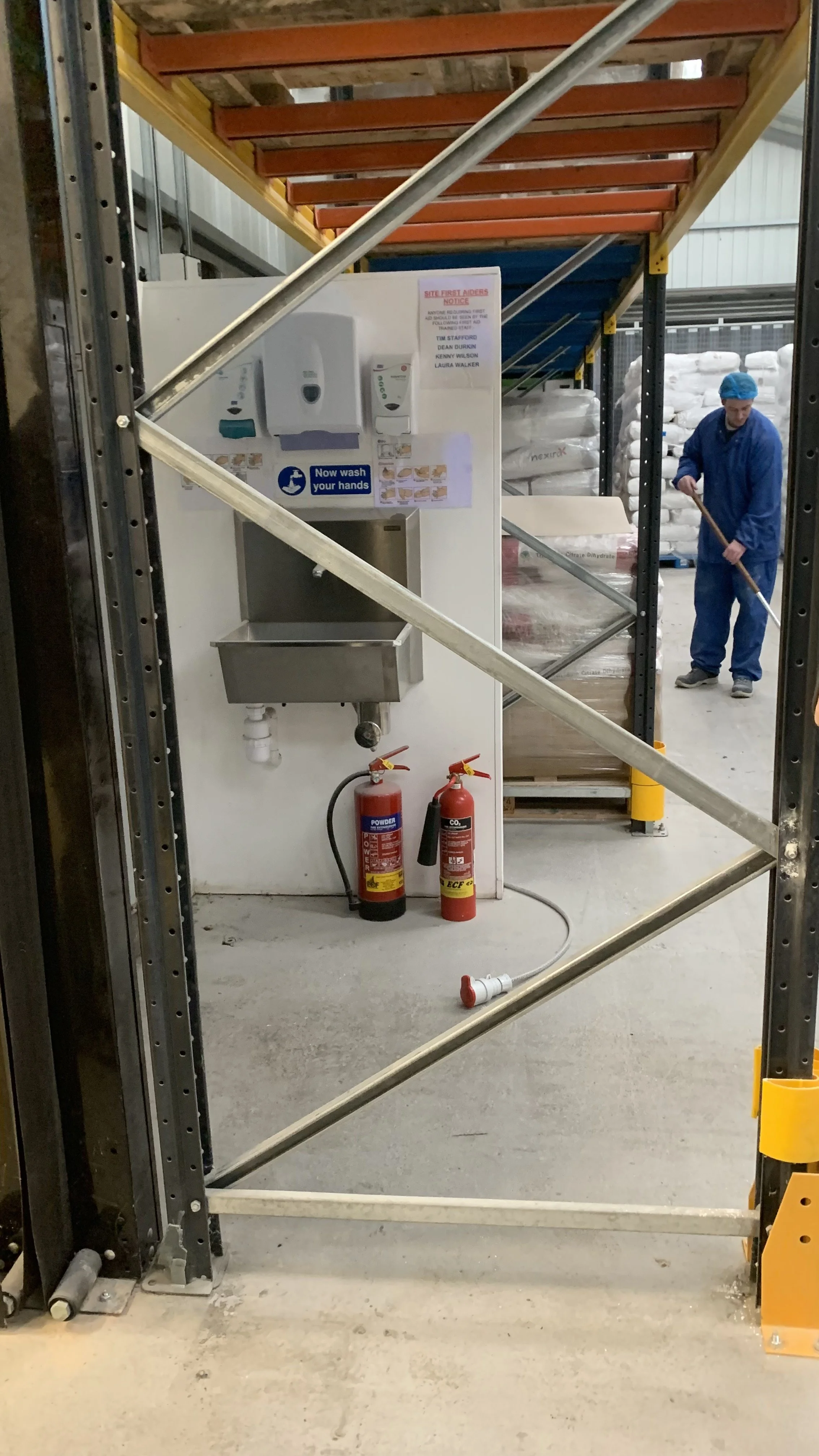 Industrial warehouse with a site first aiders notice, a handwashing station, and fire extinguishers, with a worker cleaning the floor in the background.