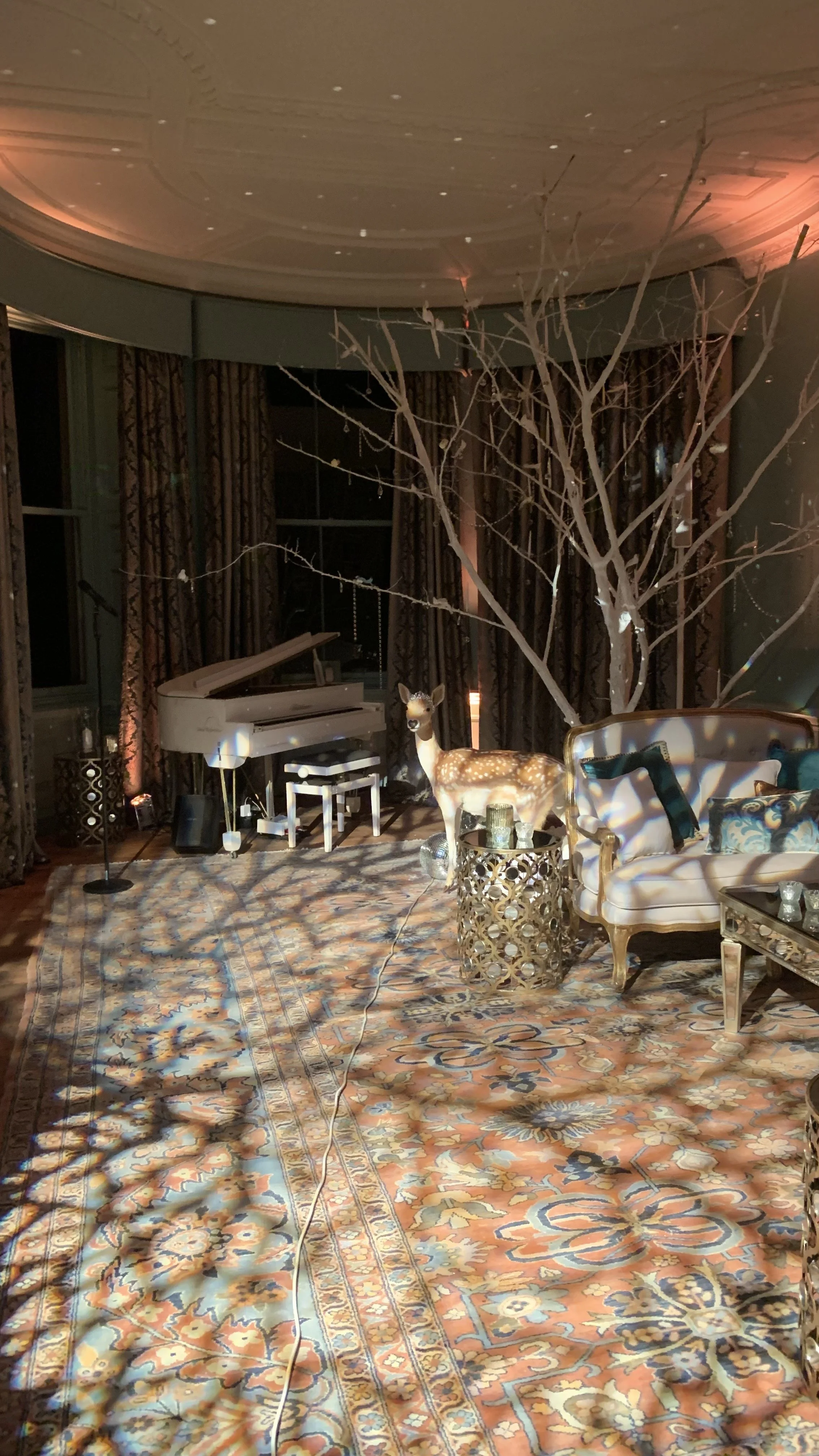 A decorated room with a white grand piano, a small stool, a tree with string lights, a deer sculpture, a gold ornate side table, a vintage sofa with pillows, a patterned rug, and a microphone stand, illuminated with colorful lighting.