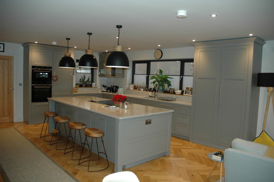 Modern kitchen with gray cabinets, marble countertops, and a large central island with three wooden barstools. Black pendant lights hang above the island, and the room has wooden flooring. There is a sink under the window, decorative items, and a pla