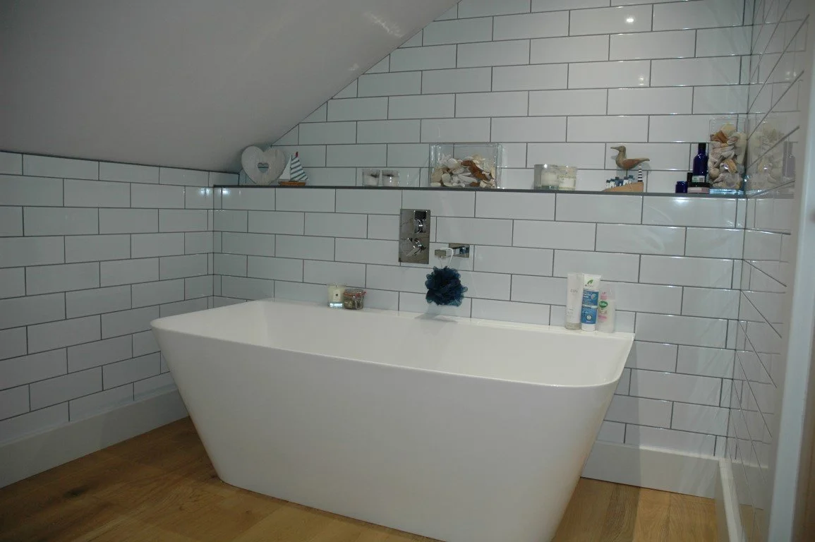 A modern bathroom with a white freestanding bathtub, white tiled walls with horizontal black grout lines, and a wooden floor. There are decorative objects and toiletries on a shelf above the tub.