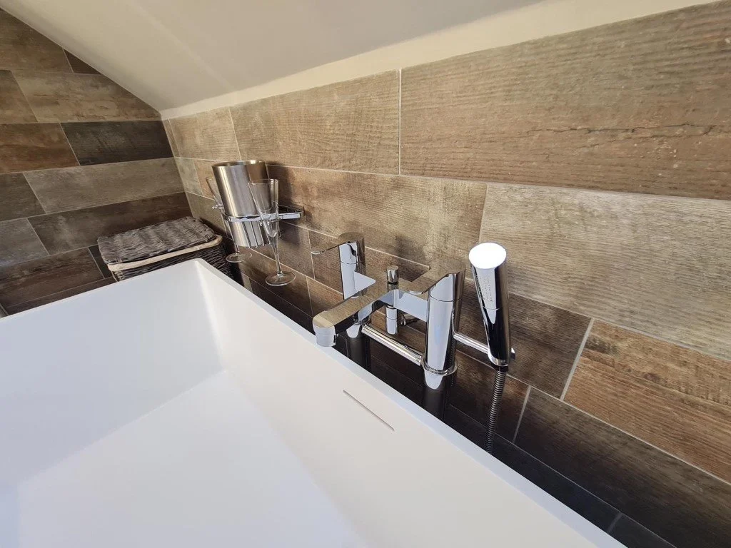 Close-up of a modern bathtub faucet with a handheld shower attachment, set against a wall with brown textured tiles. A small shelf holds metallic cups and a neatly folded towel.