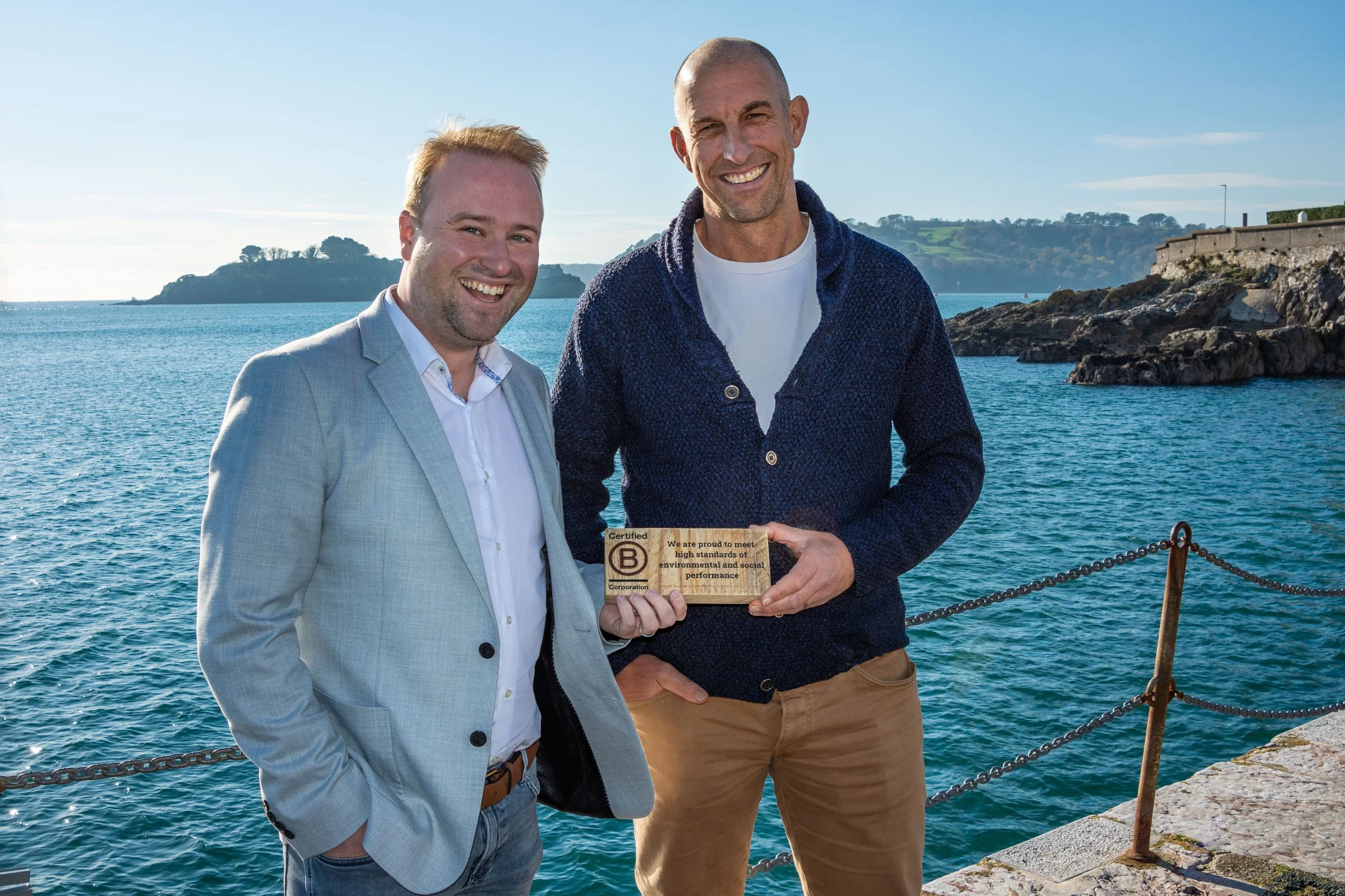 Two men stand by the water, smiling, with one holding a wooden plaque that says they meet environmental and social standards, near a rocky shore with a distant island and clear blue sky.