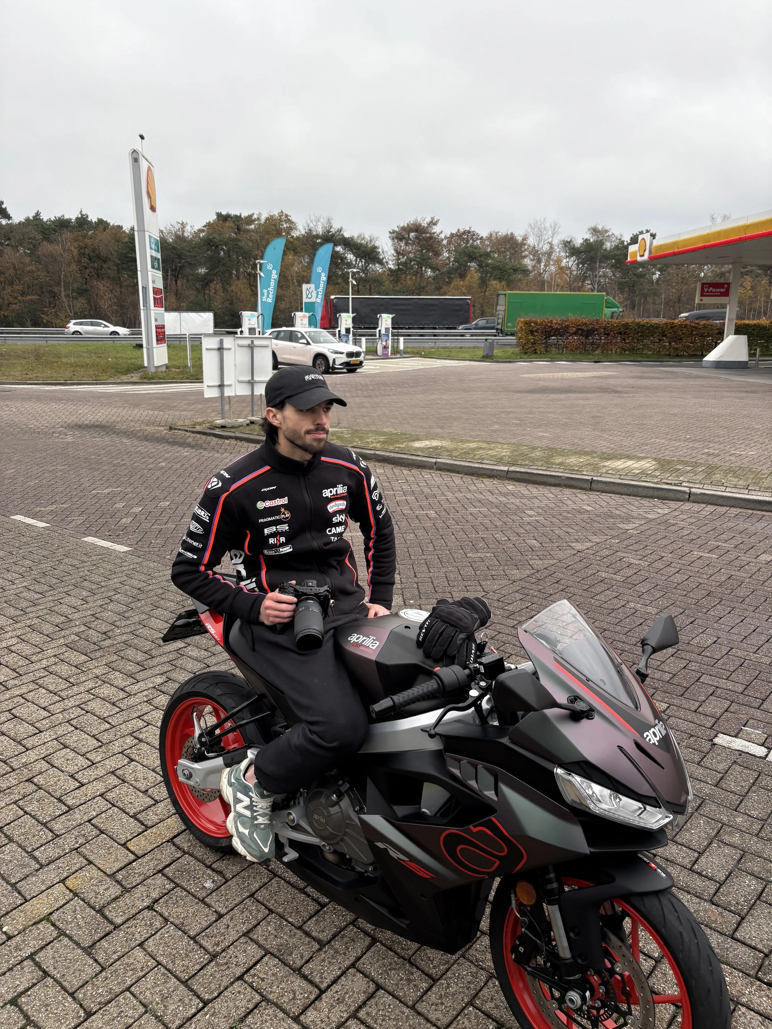 A man dressed in black racing gear sitting on a black and red motorcycle at a fuel station with electric vehicle charging stations in the background.