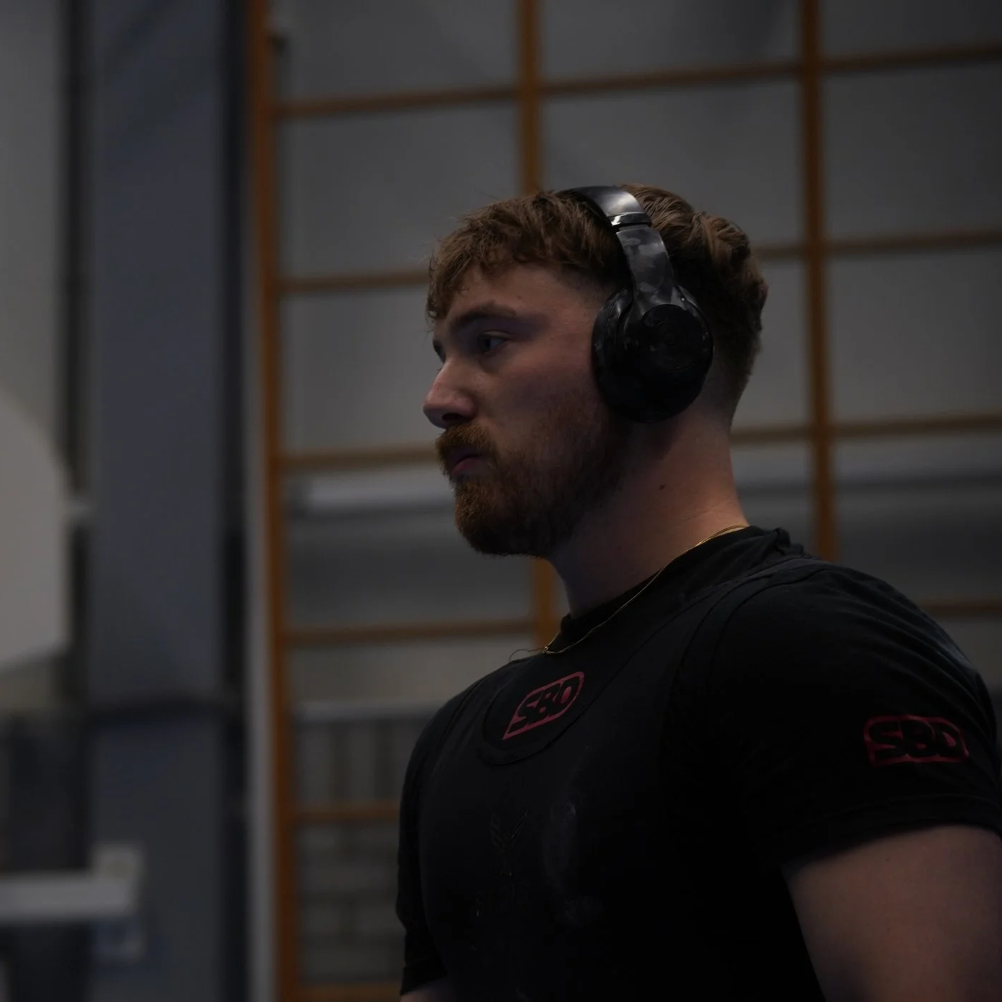 A man with a beard and curly hair wearing large black headphones and a black T-shirt with red logos, in an indoor gym or workout space.