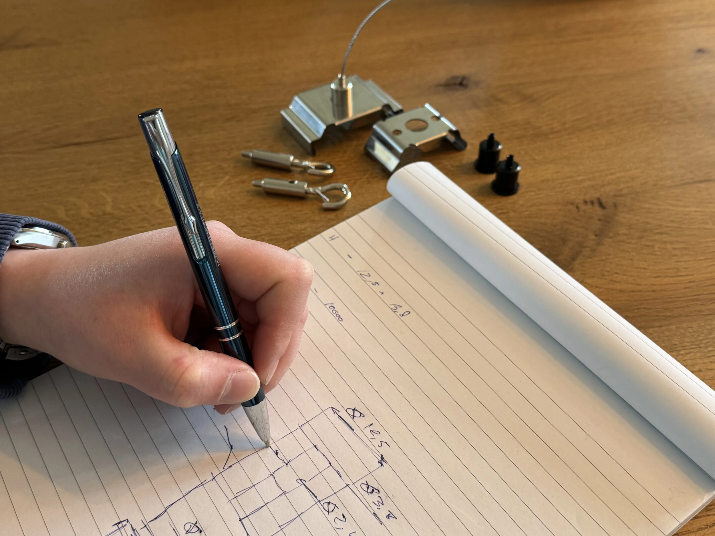 Close-up van een hand die in een notitieboekje schrijft met een balpen. Op de achtergrond liggen metalen objecten op een houten tafel.