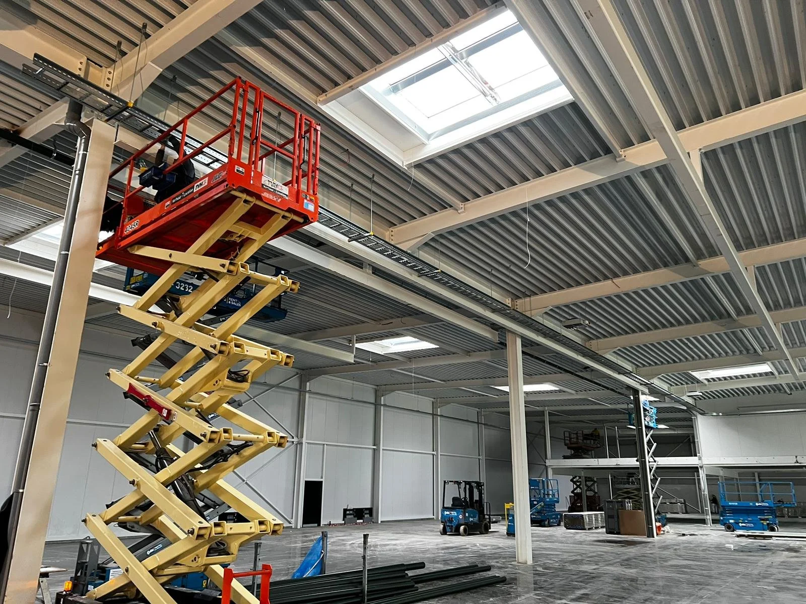 Binnen in een groot warehouse of fabriek met stellingen en apparatuur, er zijn hoogwerkers en een plafondraam dat daglicht doorlaat