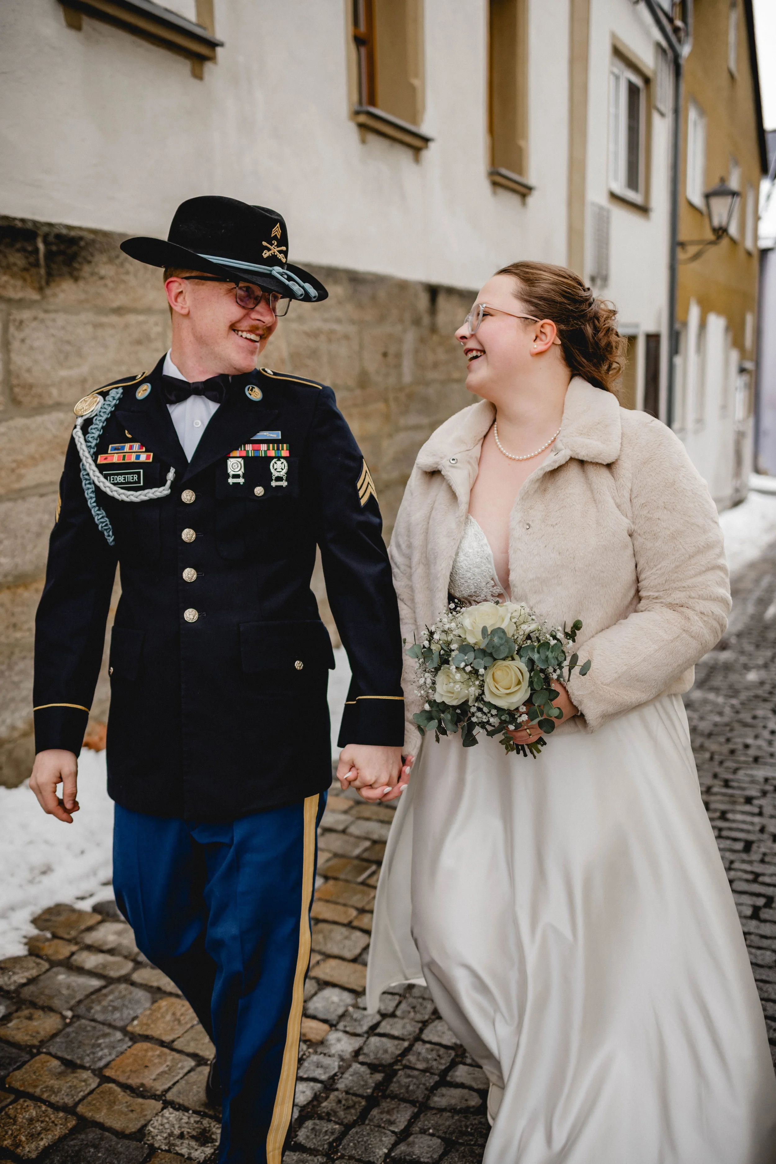 Ein glückliches Paar auf dem Hochzeitsspaziergang, die Braut trägt ein cremefarbenes Hochzeitskleid und eine Felljacke, der Bräutigam trägt eine schwarze Militäruniform. Sie halten Händchen und lachen zusammen, die Braut hat einen Blumenstrauß in der