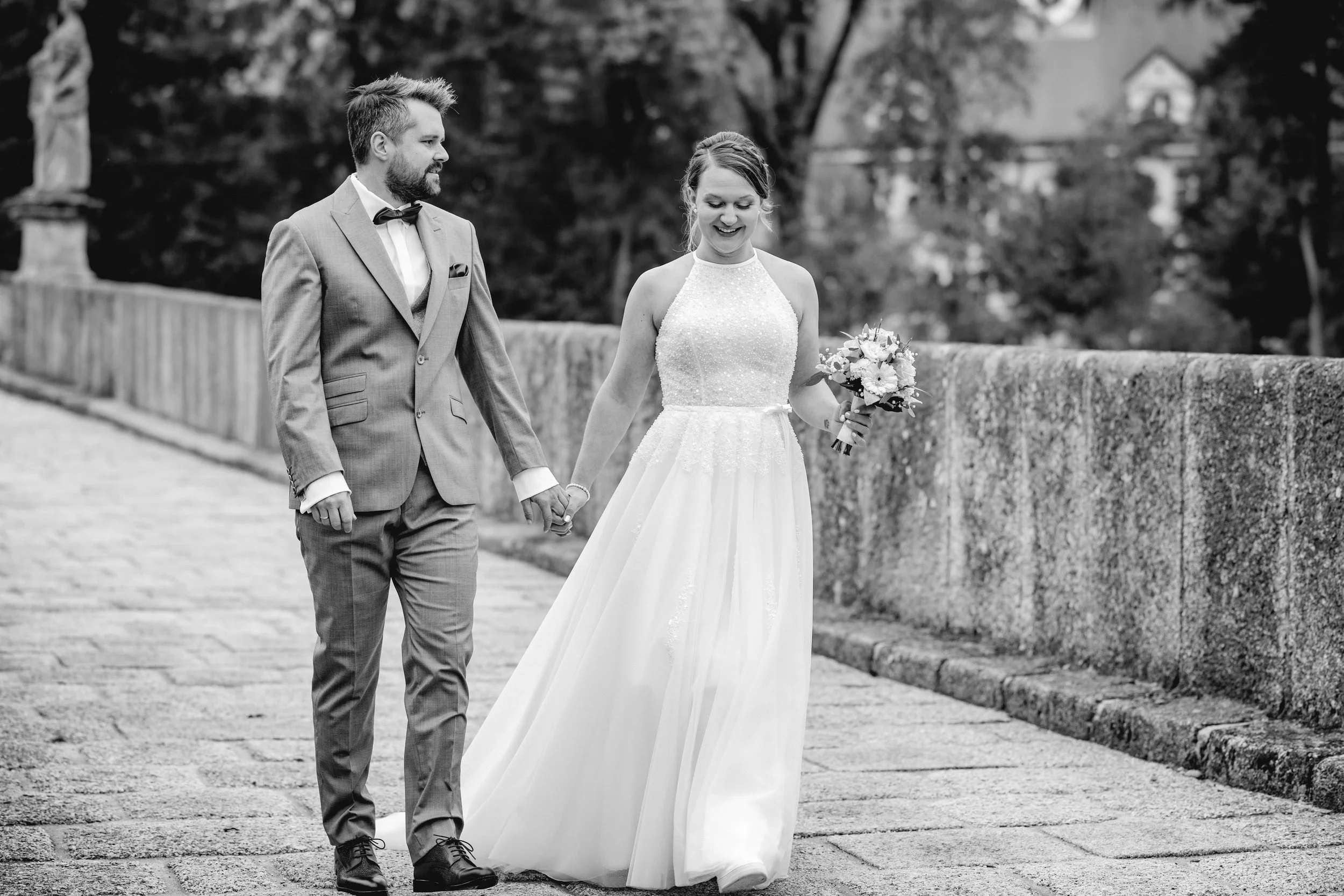 Ein Brautpaar hält Händchen beim Spaziergang im Freien, die Frau trägt ein Hochzeitskleid und hält einen Blumenstrauß, der Mann trägt einen Anzug.