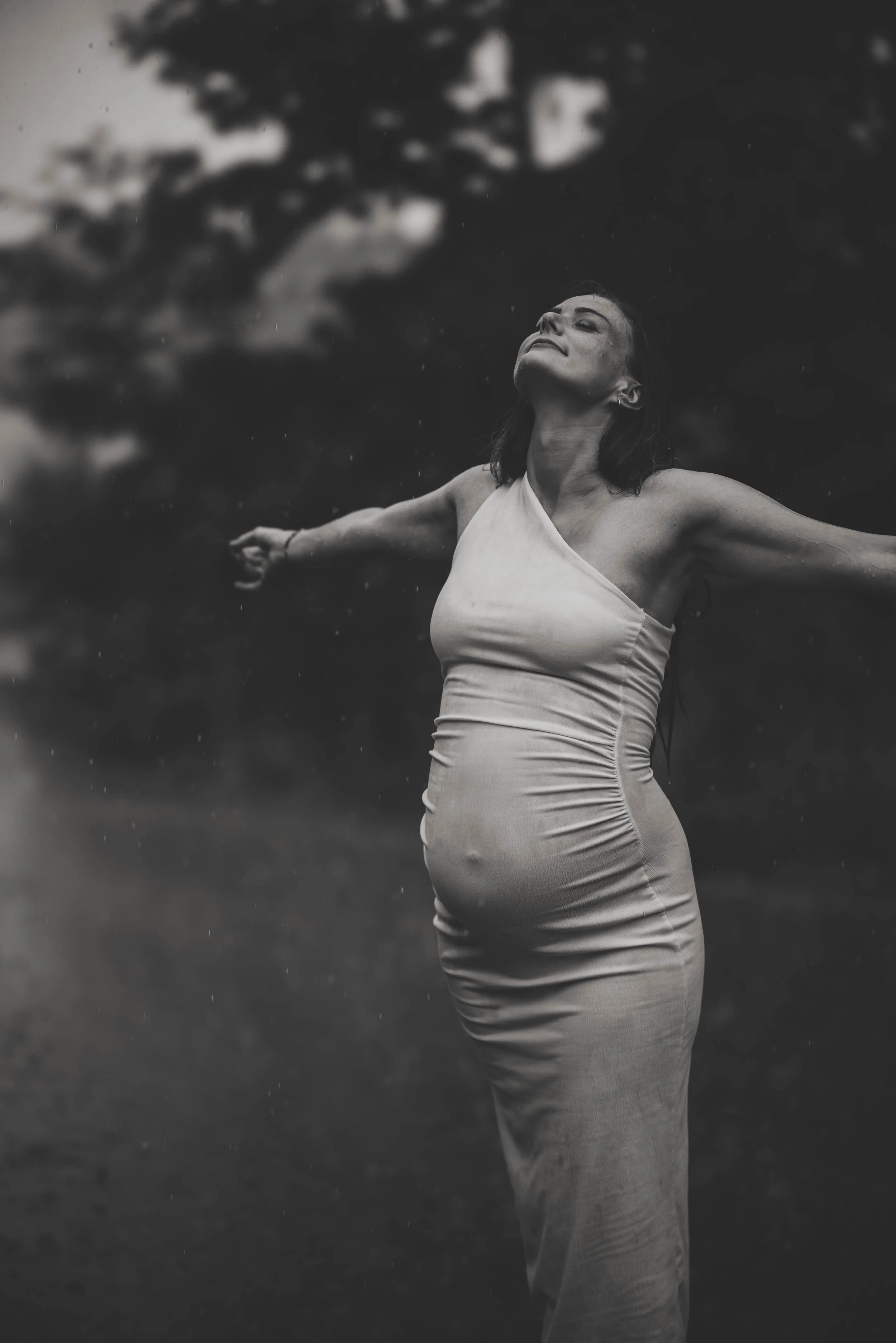 Schwarz-weiße Fotografie einer Frau mit geschlossenen Augen und ausgestreckten Armen im Freien, mit dunklem Himmel und Baum im Hintergrund.