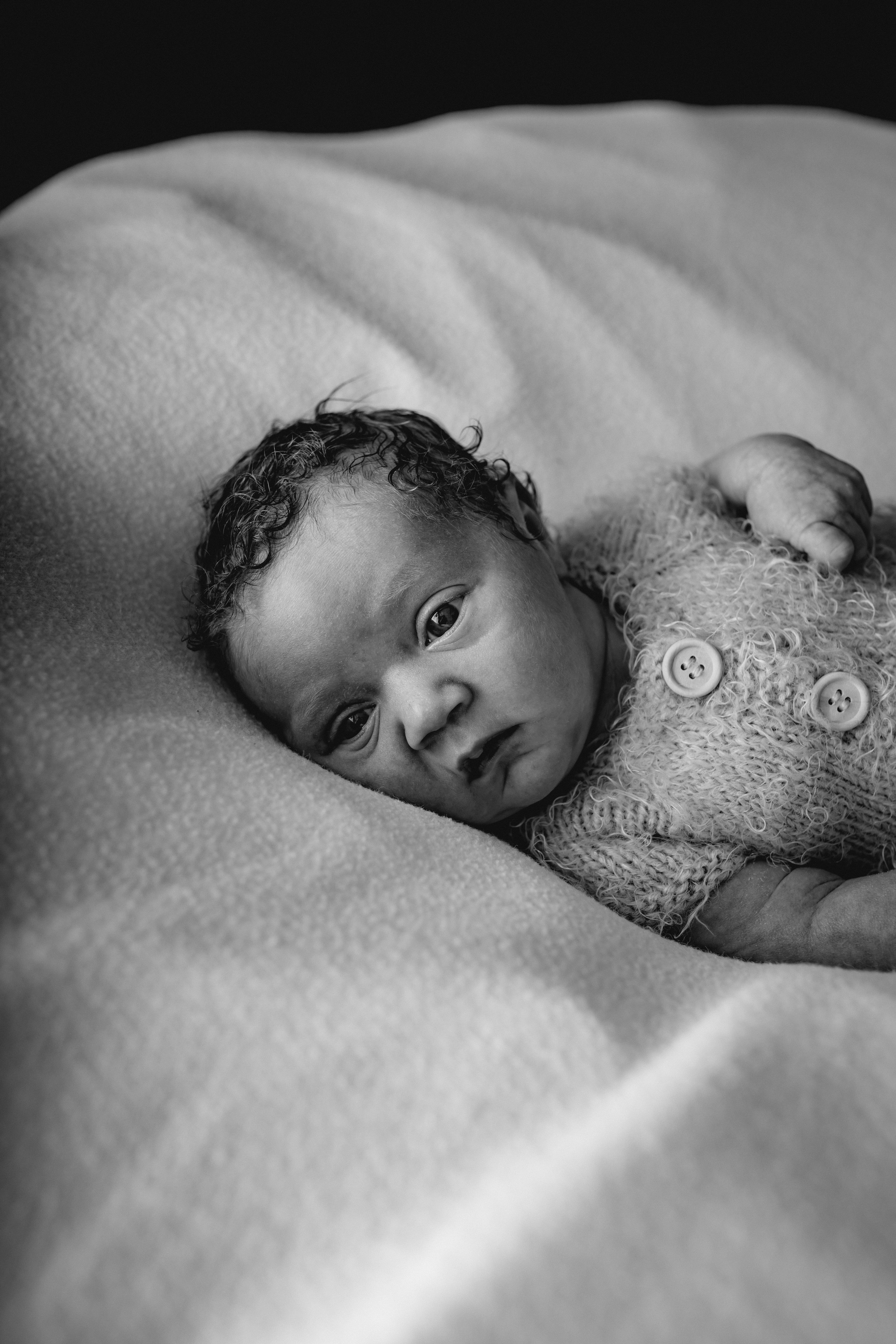 Schwarz-Weiß-Fotografie eines Babys, das auf einem weichen, großen Kissen liegt und direkt die Kamera ansieht.