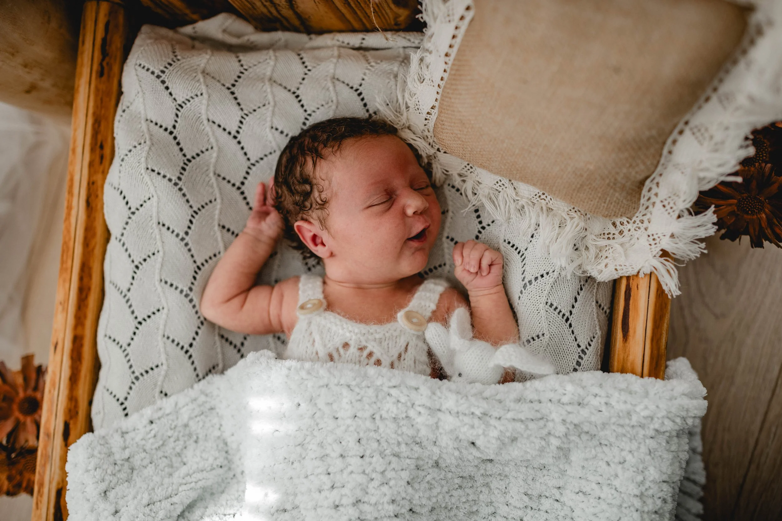 Ein schlafender Säugling liegt in einer Babywiege, umgeben von weißen Decken und einem Garnhintergrund, mit entspanntem Gesicht und verschränkten kleinen Fäusten.