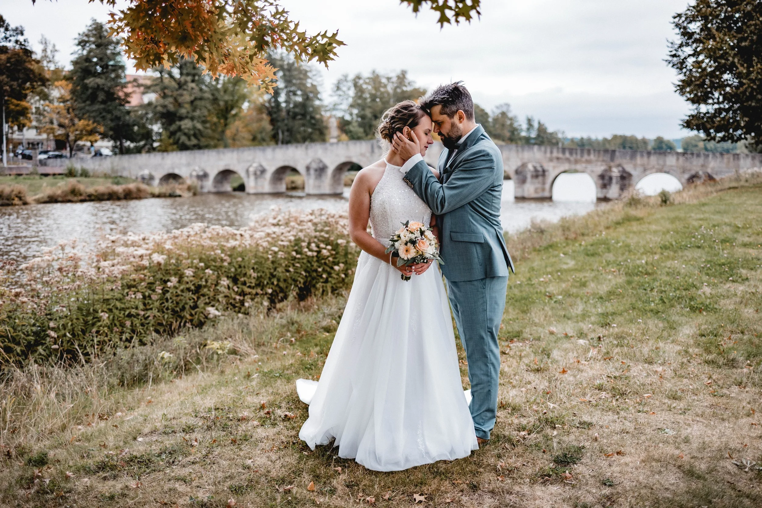 Ein heiratendes Paar steht sich nahe am Flussufer gegenüber, umarmt sich, mit einem alten Brückenbogen im Hintergrund und herbstlichen Bäumen
