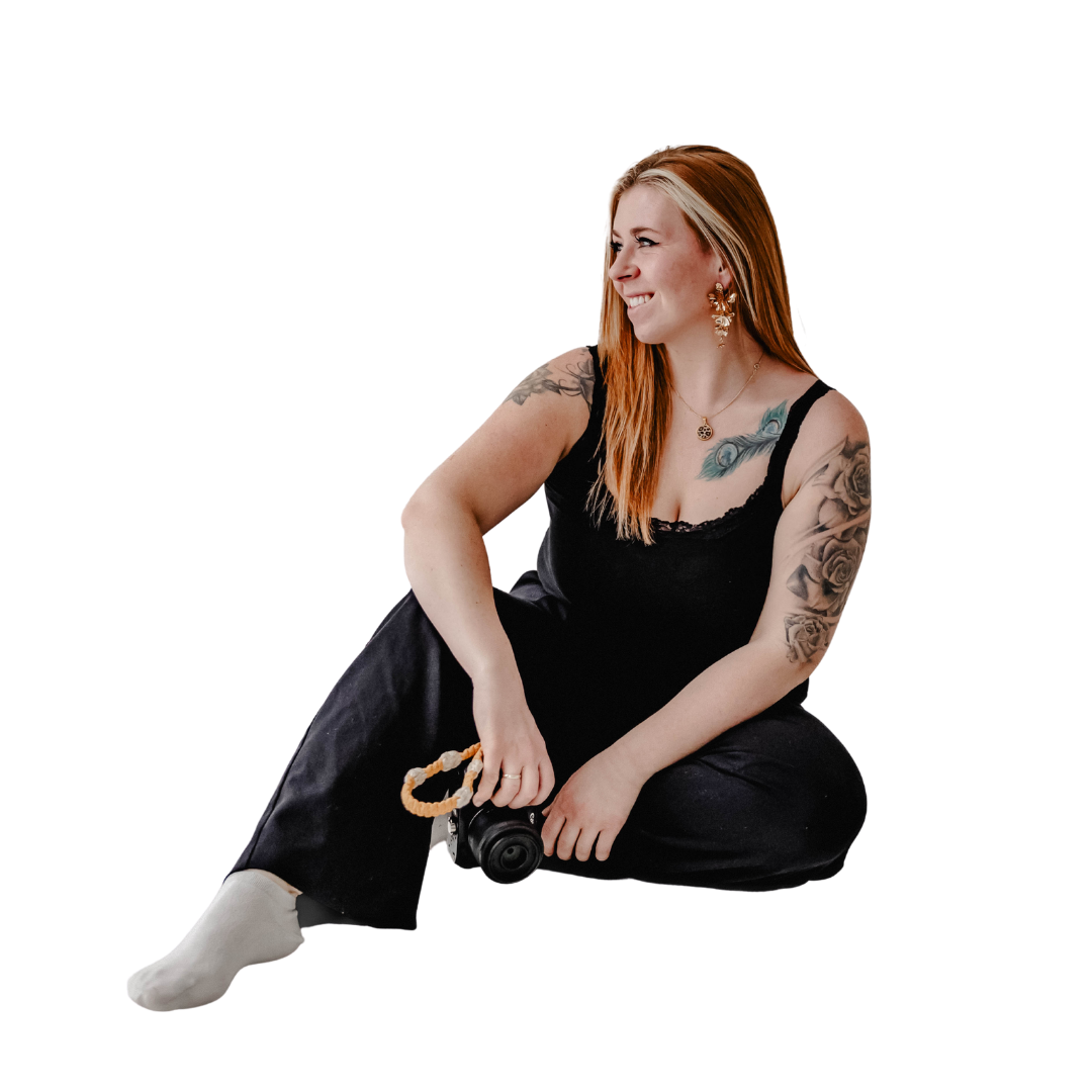 Eine lächelnde Frau mit roten Haaren, Tattoos auf Armen und Schultern, trägt schwarze Kleidung und sitzt auf dem Boden, hält eine Kamera in der Hand.