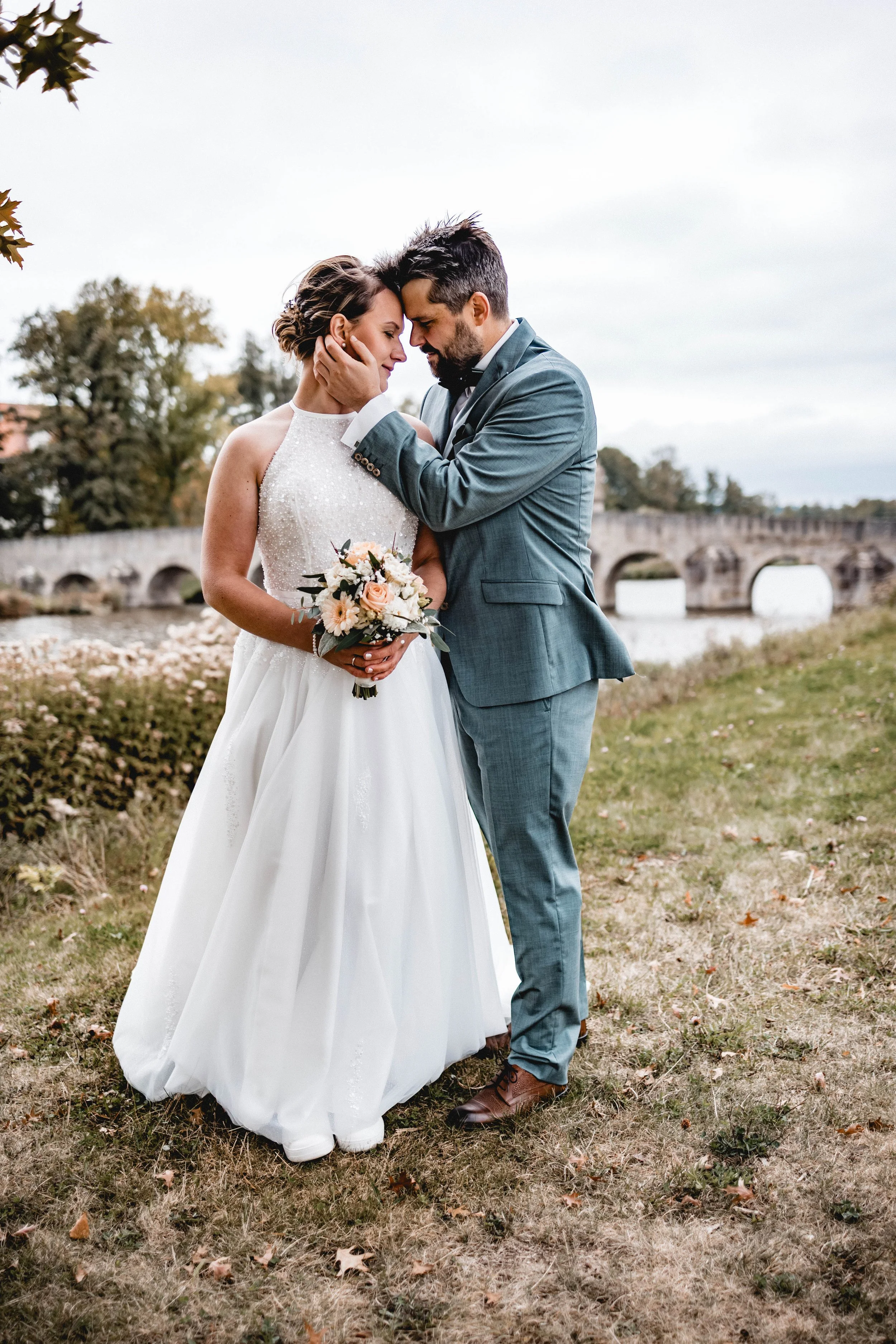 Ein Brautpaar steht eng beieinander im Freien, die Braut trägt ein weißes Hochzeitskleid, hält einen Blumenstrauß, der Bräutigam trägt einen grauen Anzug. Im Hintergrund ist eine Brücke und ein Fluss zu sehen, das Wetter ist bewölkt.