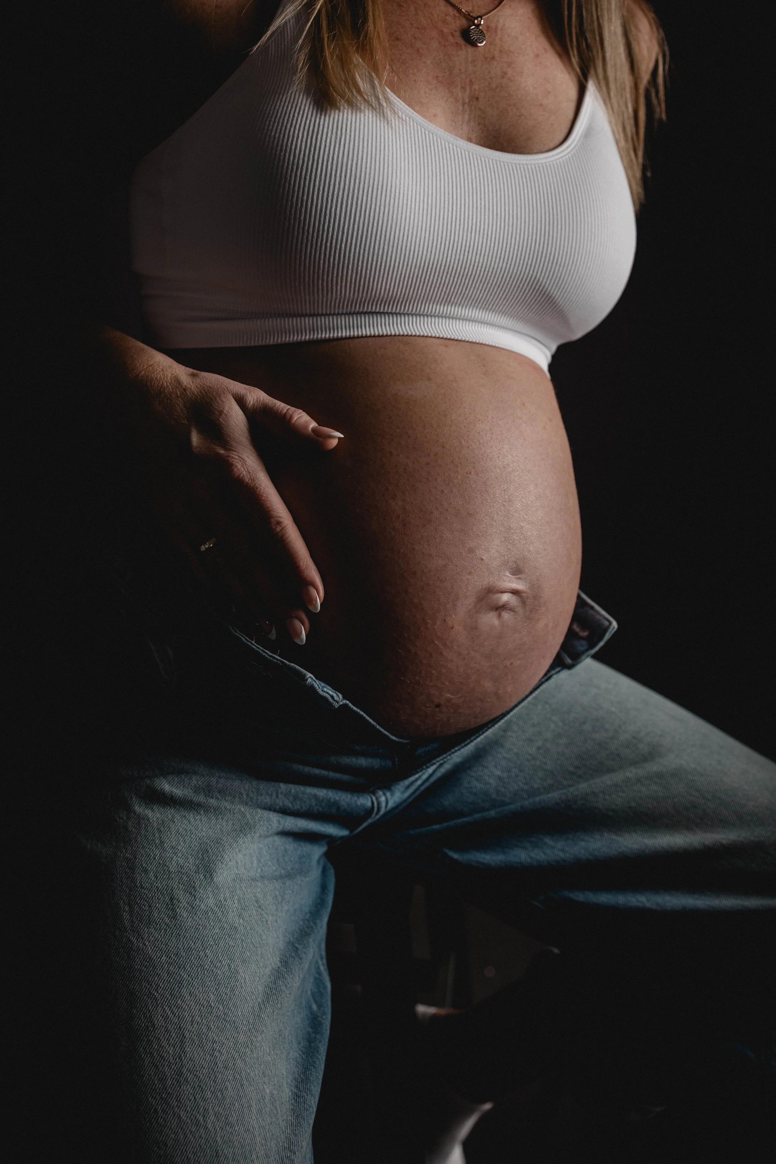 Schwangere Frau, die ihren Bauch hält, trägt ein weißes Oberteil und sitzt auf einem Stuhl gegen einen dunklen Hintergrund.