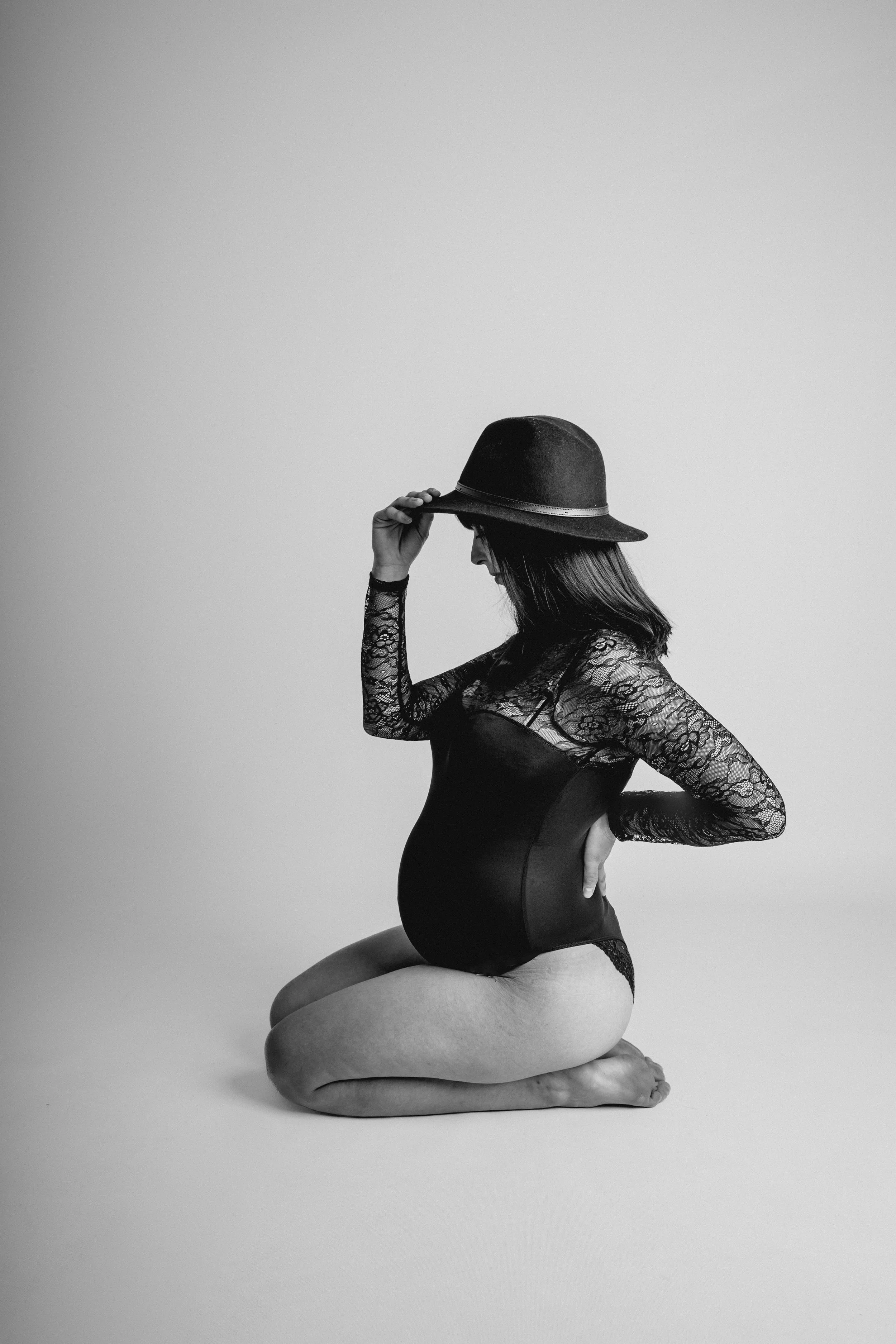 Schwangere Frau in schwarzen Spitzenkleid und Hut, sitzend auf Knien in Schwarz-Weiß-Fotografie.