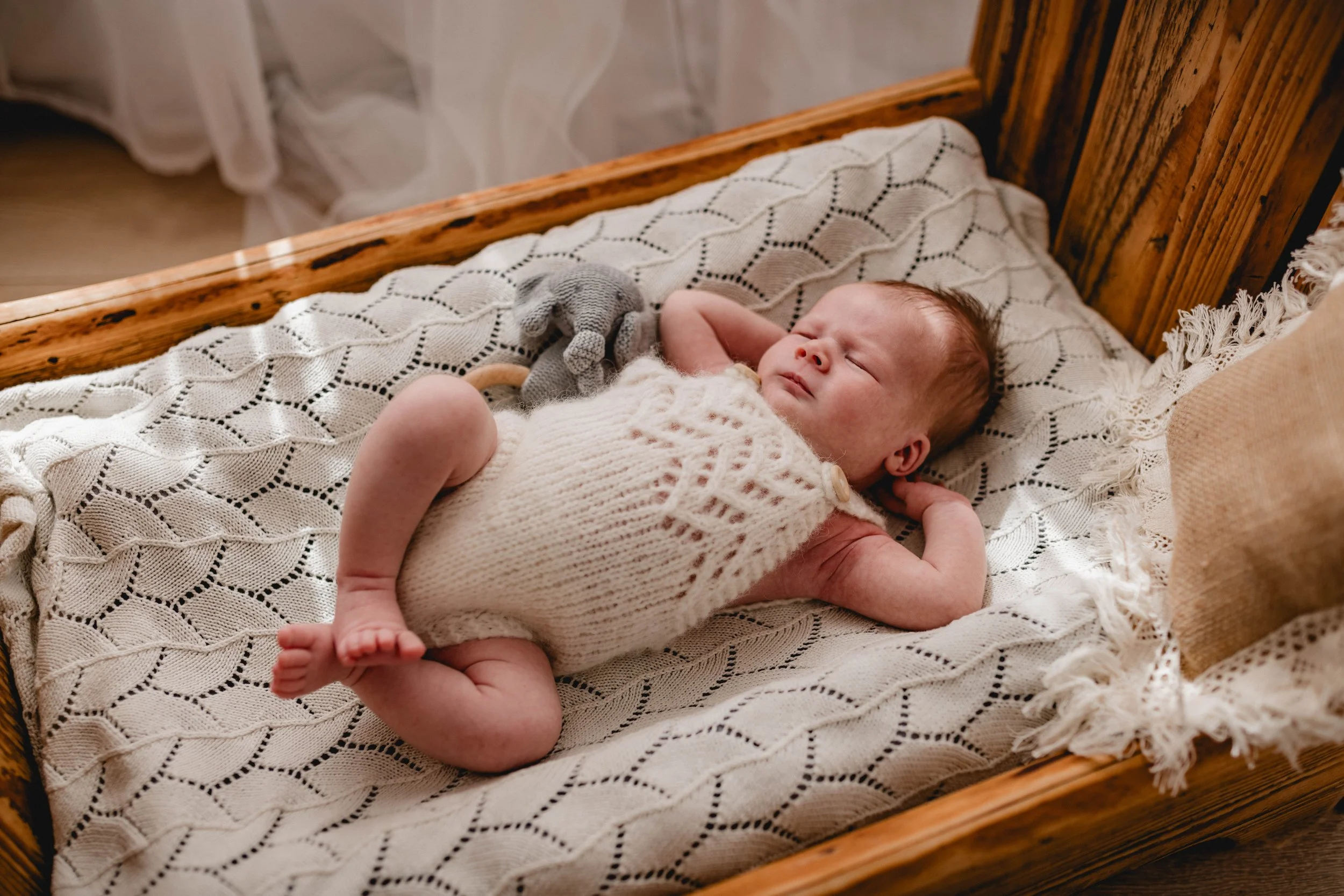 Ein neugeborenes Baby liegt in einem Holzbett auf einem Decken auf einer Matratze, trägt einen weißen Strickoverall und hält eine graue Stoffpuppe. Das Baby schläft oder ist im Begriff einzuschlafen.