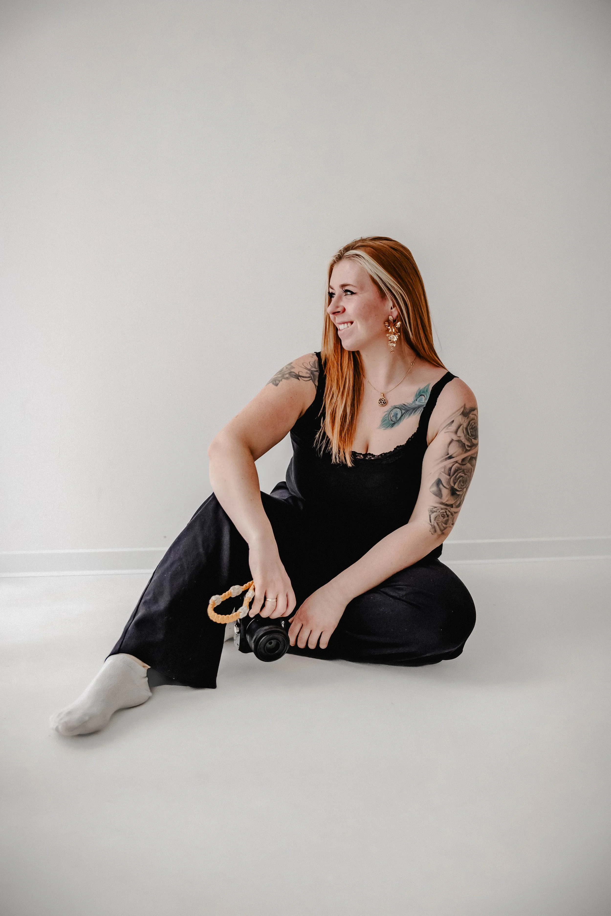Eine Frau mit roten Haaren, Tattoos auf Armen und Schultern, trägt eine schwarze Kleidung, sitzt auf dem Boden, hält eine Kamera in der Hand und lächelt. Die Hintergrundwand ist hell und leer.
