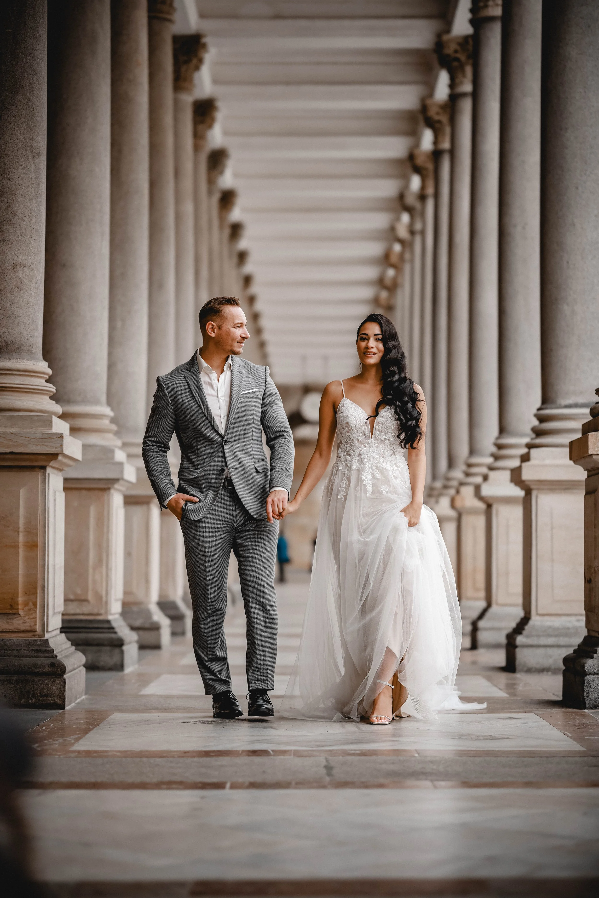 Ein Paar in Hochzeitskleidung spaziert durch eine Säulenhalle, hält Händchen und lächelt sich an.