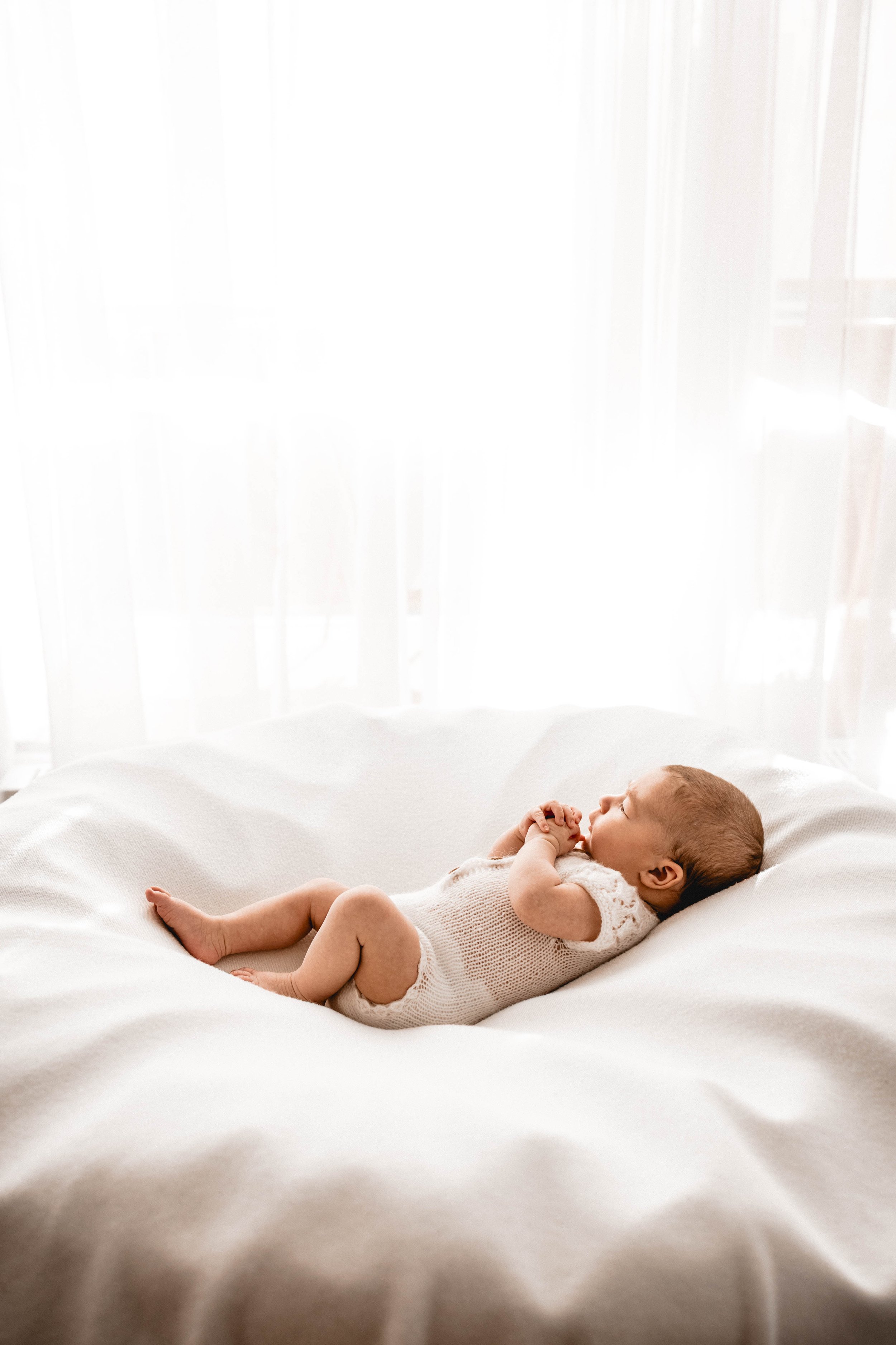 Kleiner Säugling liegt auf einem weißen Bett vor einem hellen Vorhang, schläft friedlich, trägt einen Strickbody und liegt auf dem Rücken.