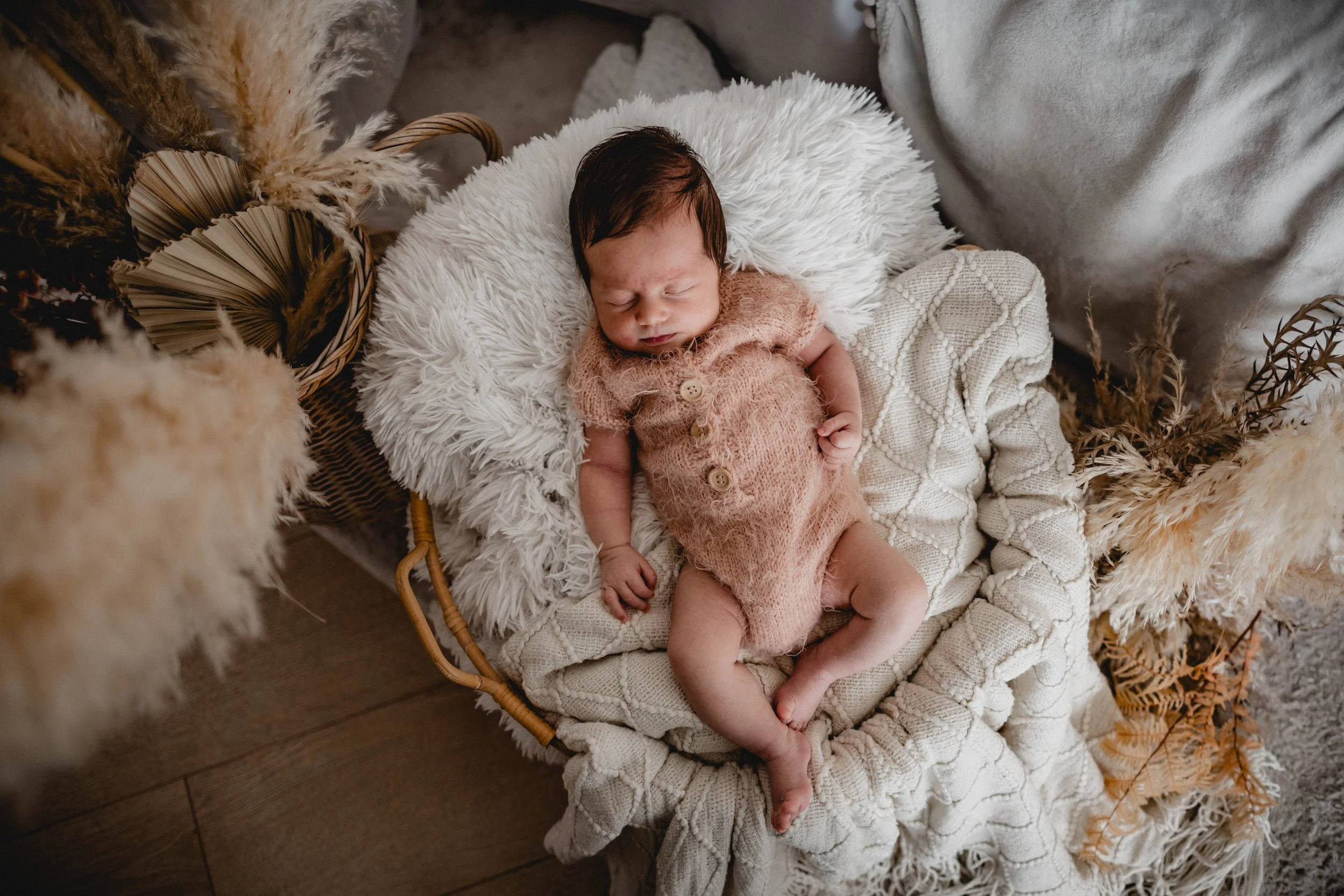 Ein schlafendes Baby in einem gestrickten, pinken Kleid liegt in einem Korb mit weichen Decken und Trockensträußen, umgeben von Dekorationen.