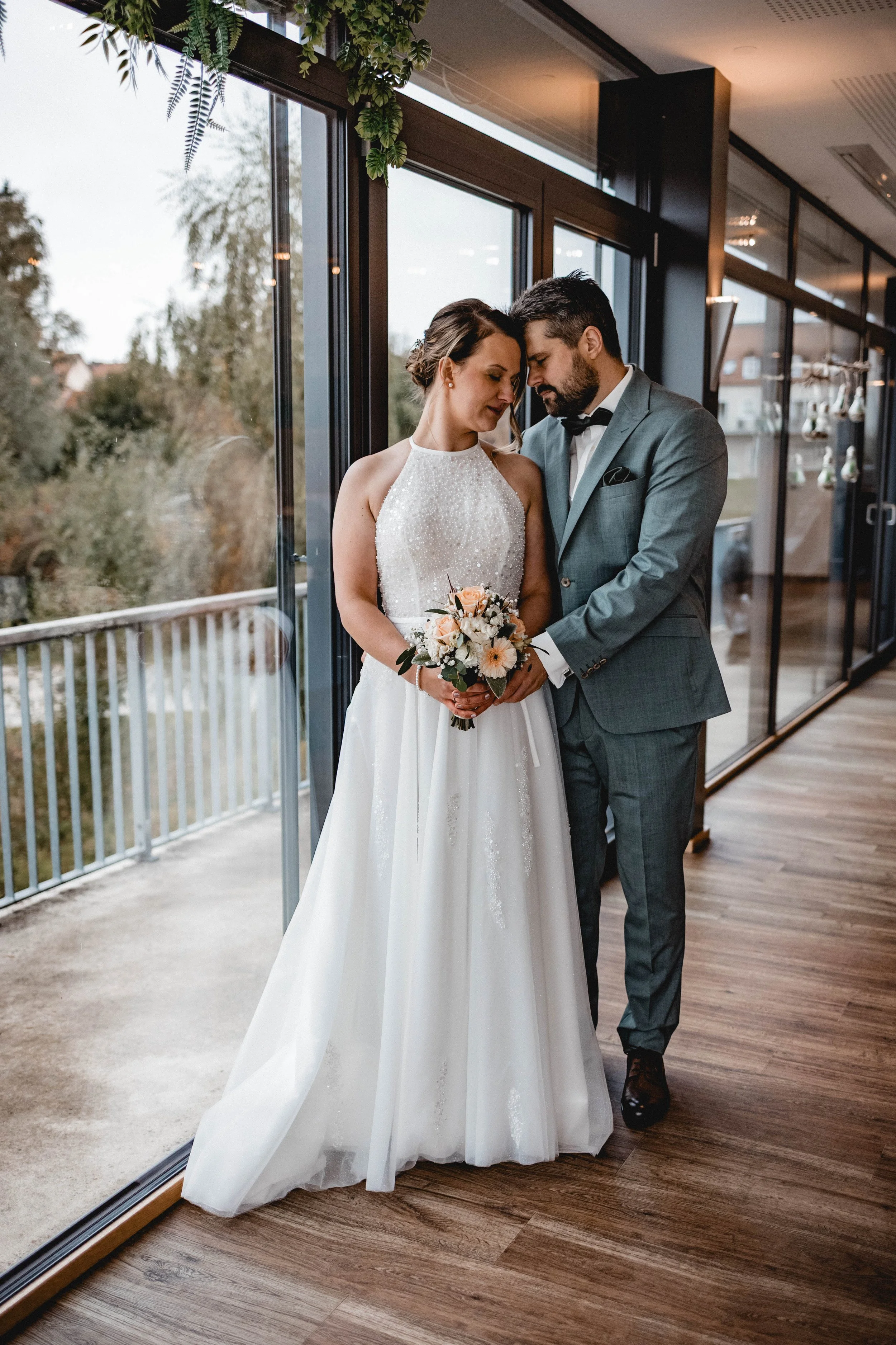 Ein Brautpaar steht drinnen vor großen Fenstern und hält sich an den Händen, die Braut trägt ein weißes Brautkleid und hält einen Blumenstrauß, der Bräutigam ist in einem grauen Anzug mit schwarzem Sakko gekleidet.